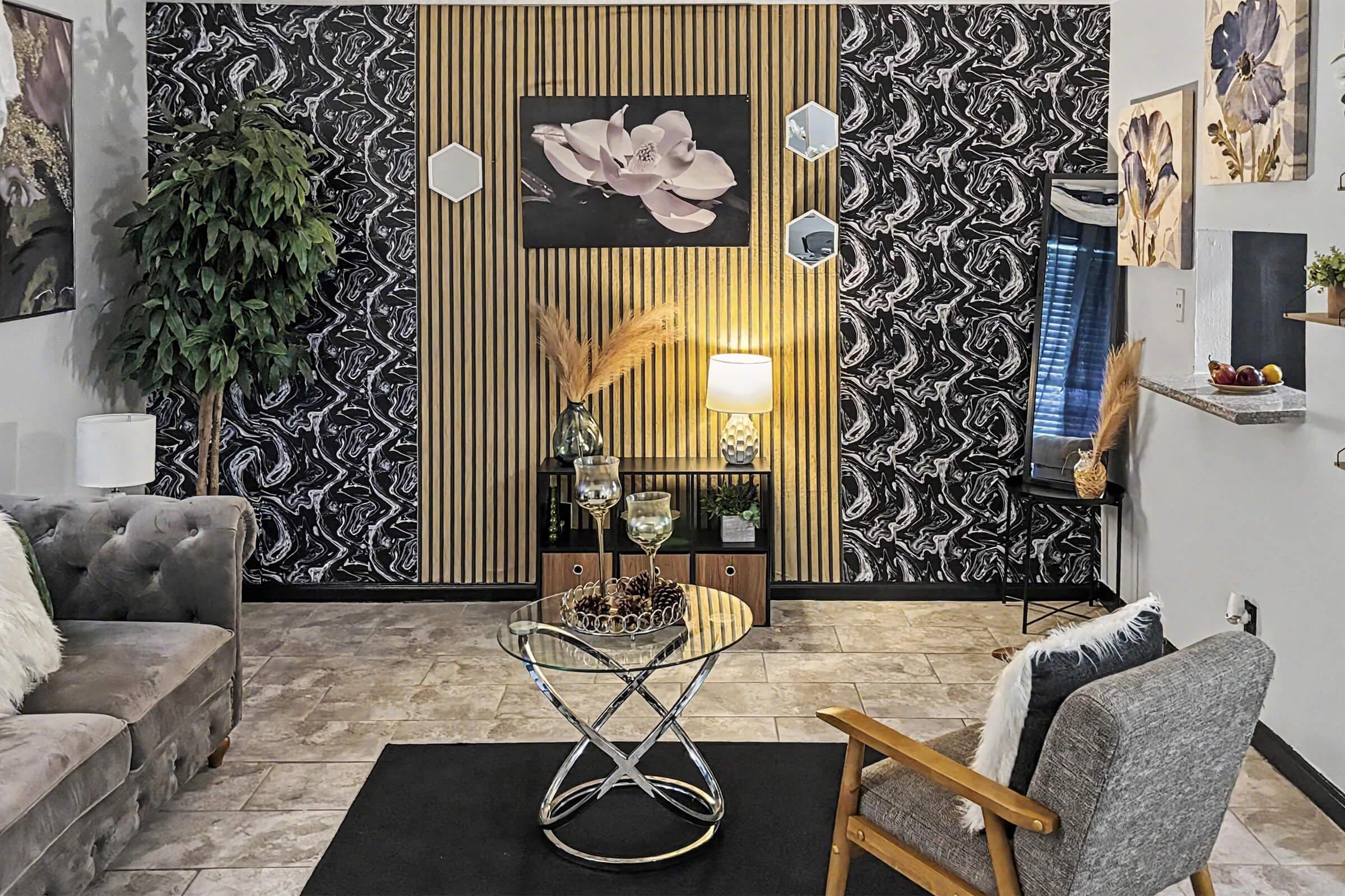 A cozy living room featuring a plush gray sofa, a glass coffee table with decorative items, and a stylish armchair. The wall behind is adorned with a bold patterned wallpaper and wooden slats, complemented by houseplants and modern decor. A warm lamp provides soft lighting, enhancing the inviting atmosphere.