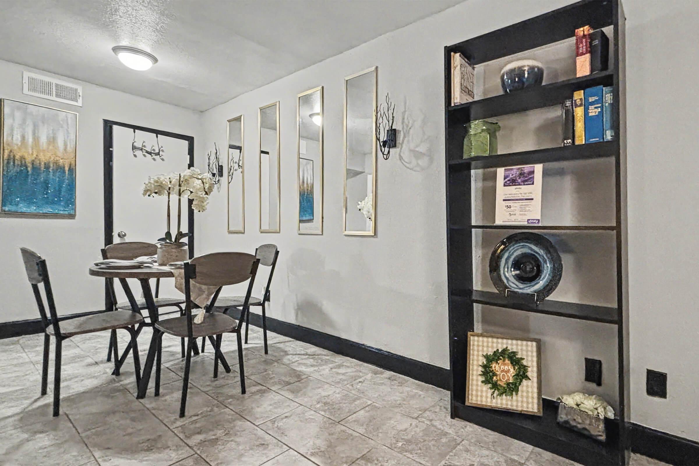 A modern dining area featuring a small round table with four chairs, alongside a black bookshelf displaying decorative items. The walls are adorned with mirrors and wall sconces, and there's a piece of abstract art in blue and gold. Neutral tile flooring adds to the contemporary aesthetic.
