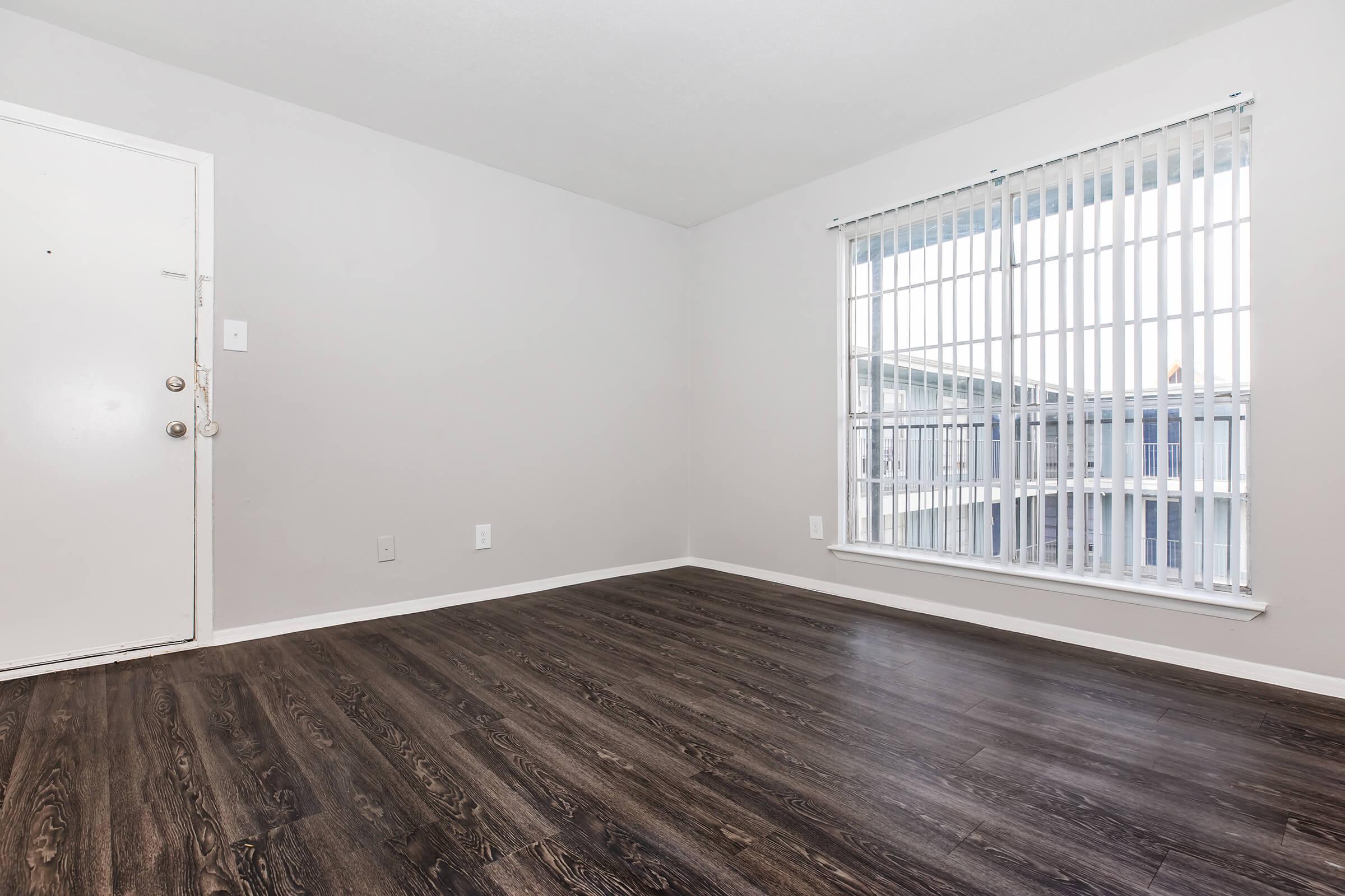 Empty room with light gray walls, a large window with white vertical blinds letting in natural light, and dark wood laminate flooring. The door is visible on the left side, and there's ample space in the room, which appears neat and unfurnished.