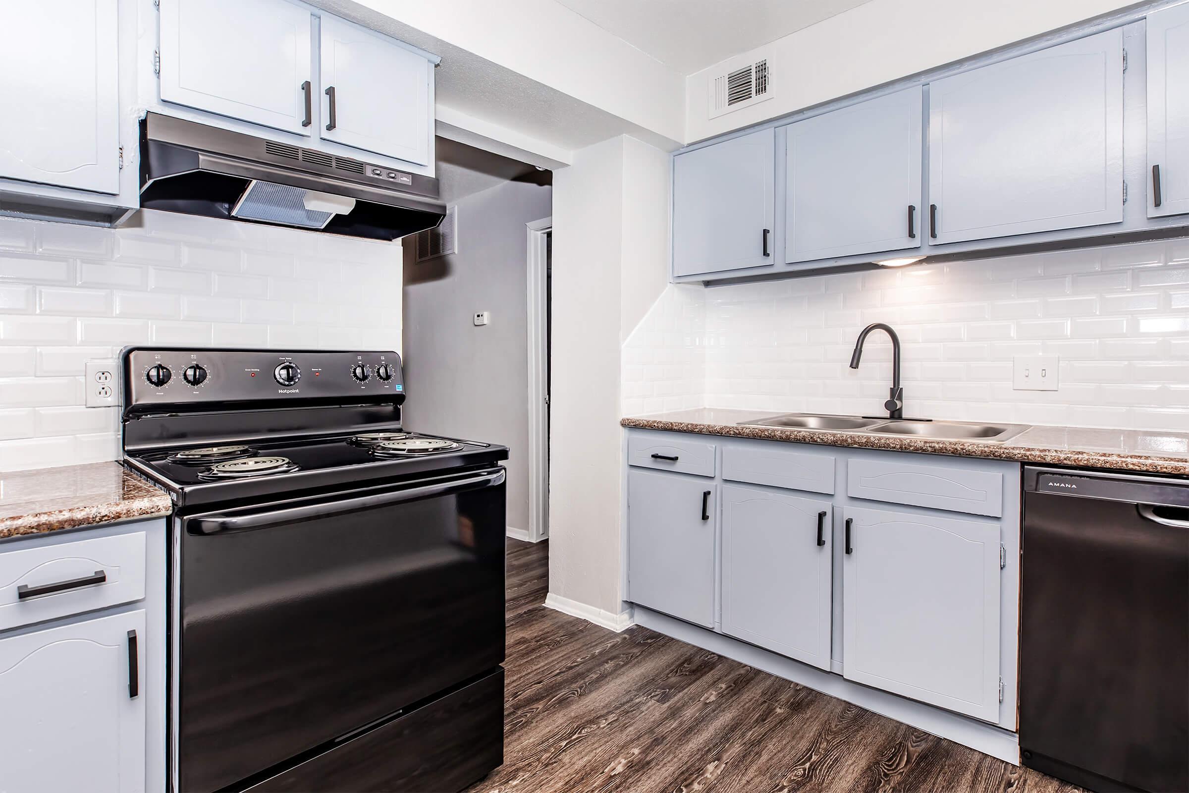 Modern kitchen featuring blue cabinets, a black stove, a stainless steel sink with a faucet, and a dark countertop. The walls are tiled with white subway tiles, and the flooring is a dark wood laminate. Overall, the space is bright and inviting with a clean design.