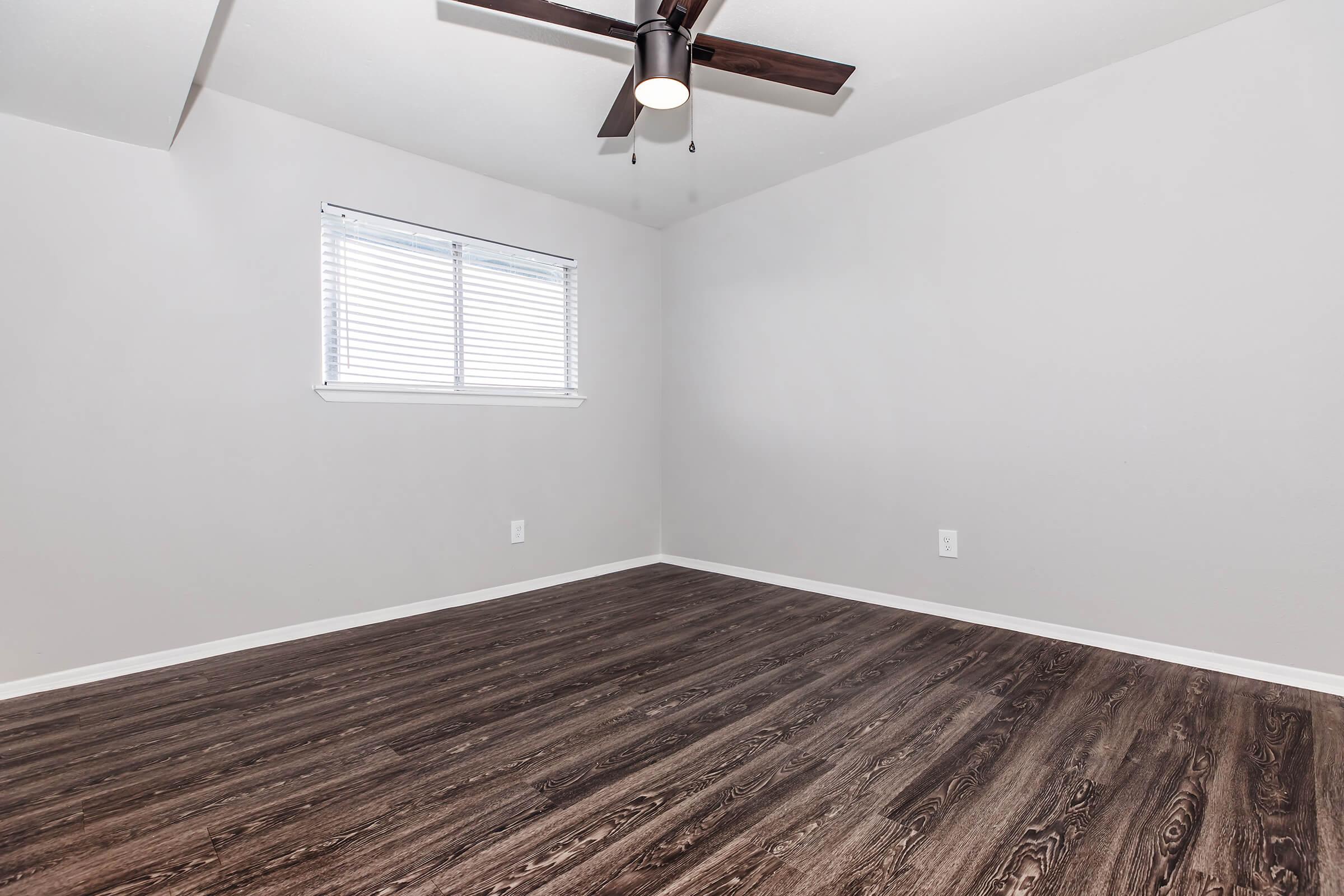 A spacious room featuring light gray walls, a ceiling fan with dark blades, and a window with white blinds. The floor is made of dark wood laminate, creating a modern aesthetic. The room is empty, emphasizing its openness and potential for personal decoration.