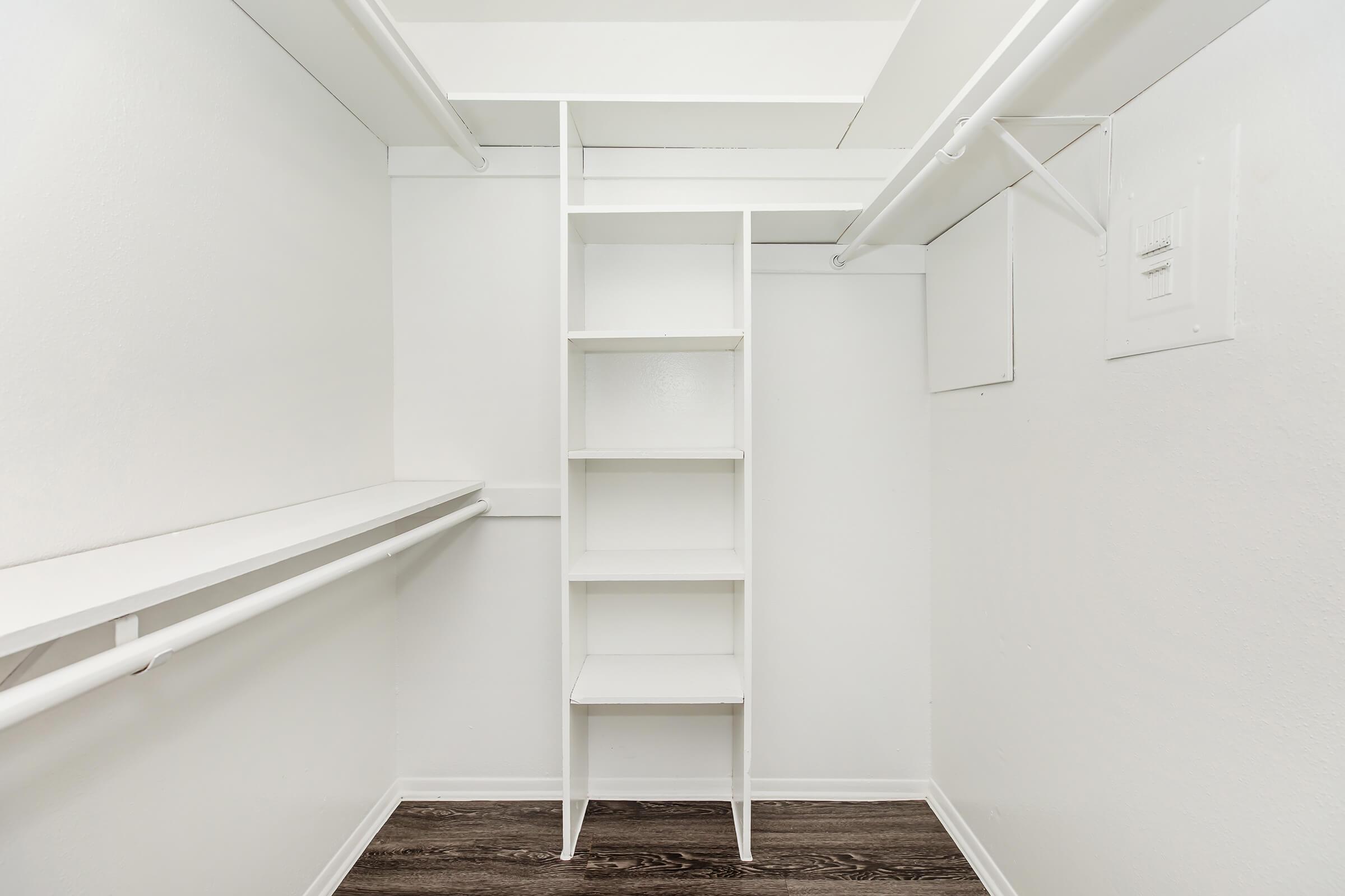 A clean, empty closet featuring white walls, a built-in shelf unit with five shelves, and a hanging rod. The floor has dark wood-like flooring, contributing to a minimalist and modern appearance.
