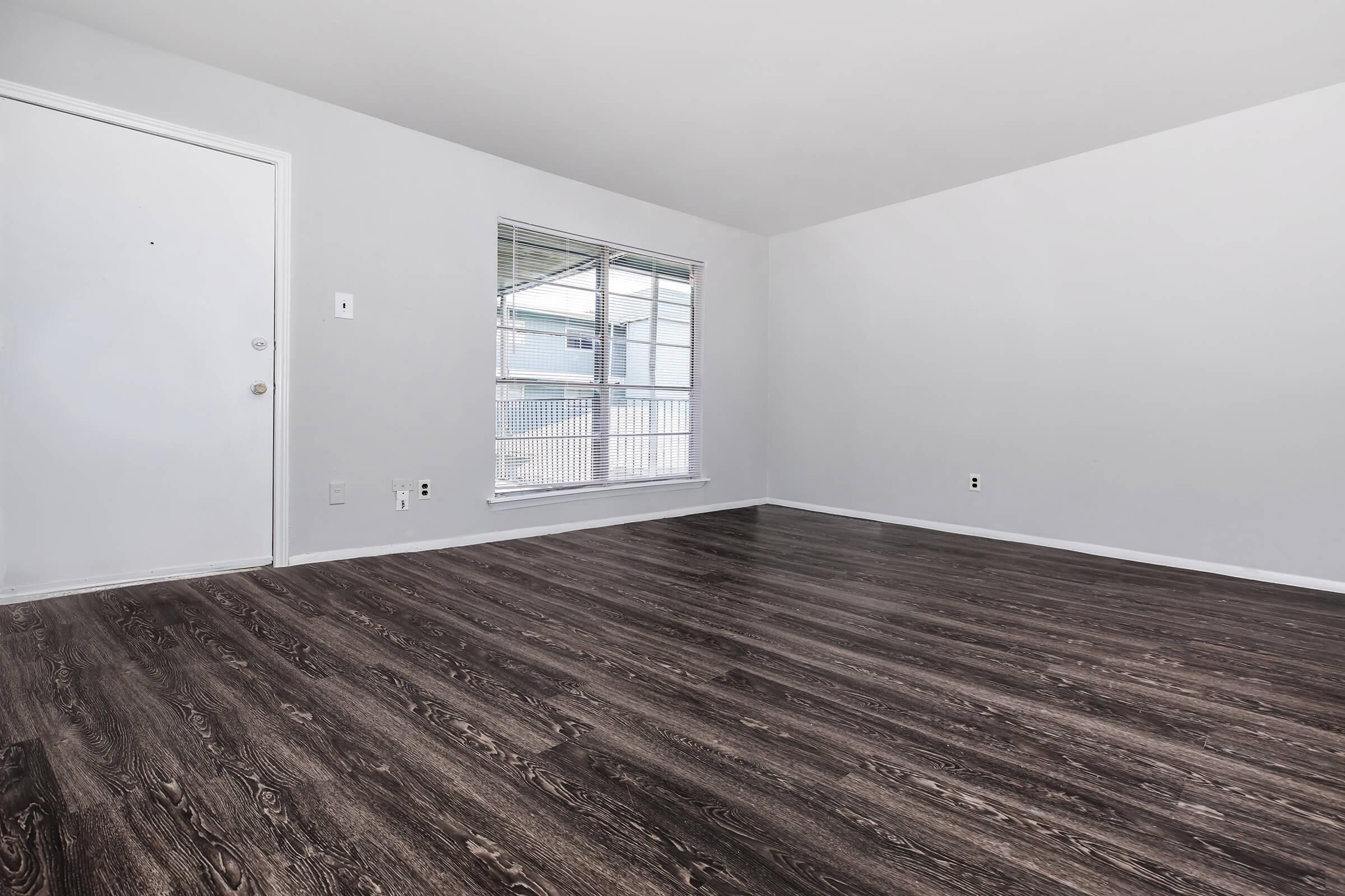 Empty room with light gray walls and dark wooden vinyl flooring. A closed white door is on the left, and a window with blinds allows natural light to enter. The space is unfurnished, creating a clean and open atmosphere.
