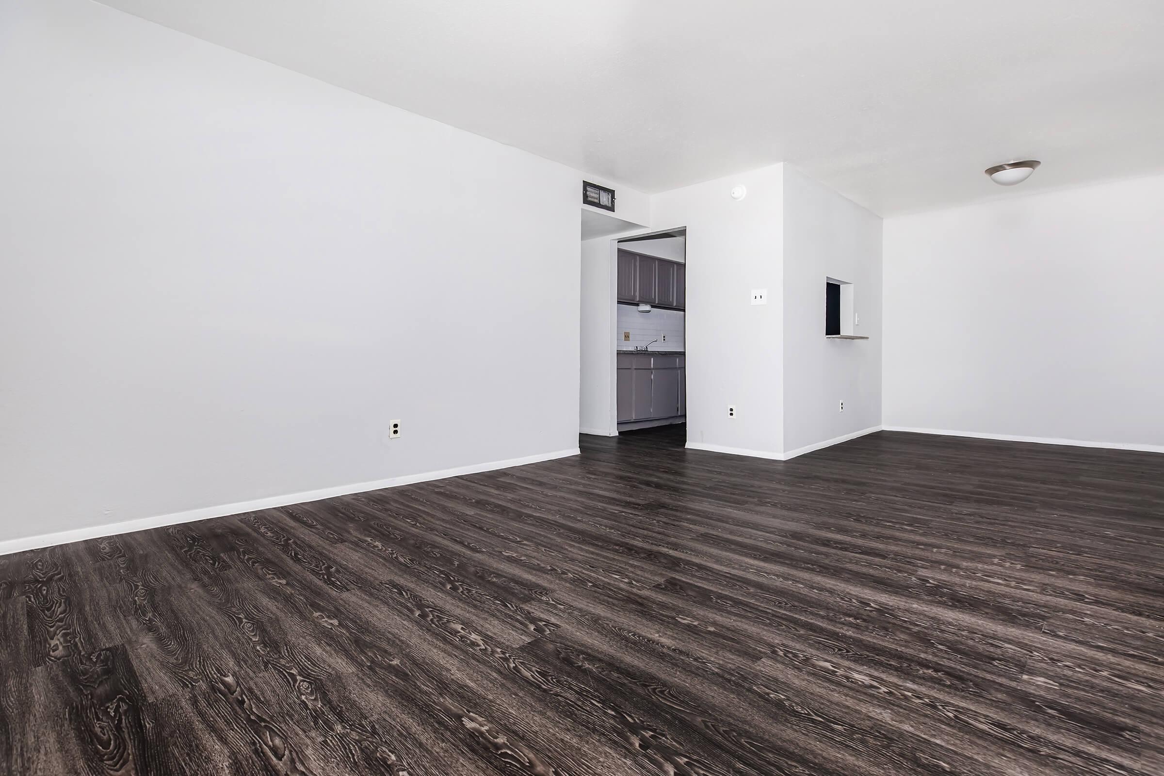 A spacious interior view of a room with dark wood-style flooring, light gray walls, and a doorway leading to a kitchen area. Natural light fills the space, creating a bright and inviting atmosphere. The layout is open with minimal furnishings, emphasizing the potential for customization.