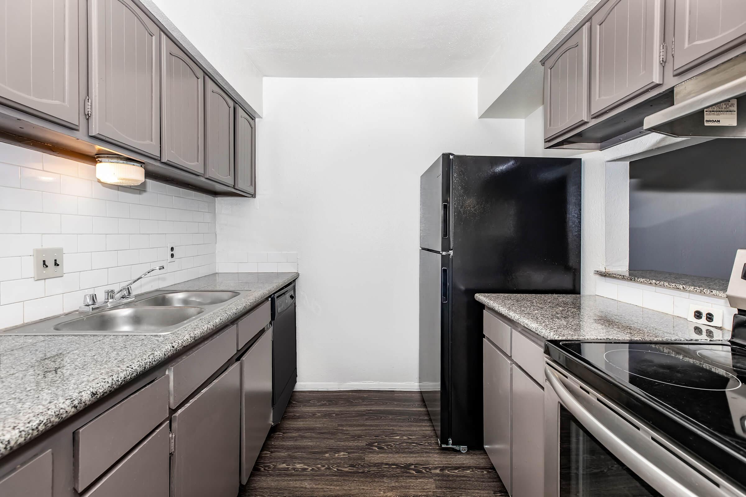 A modern kitchen featuring dark gray cabinets, white tiled backsplash, a double sink, a black refrigerator, and stainless steel appliances, including an oven and stove. The flooring is a dark wood laminate, creating a contemporary and clean atmosphere.