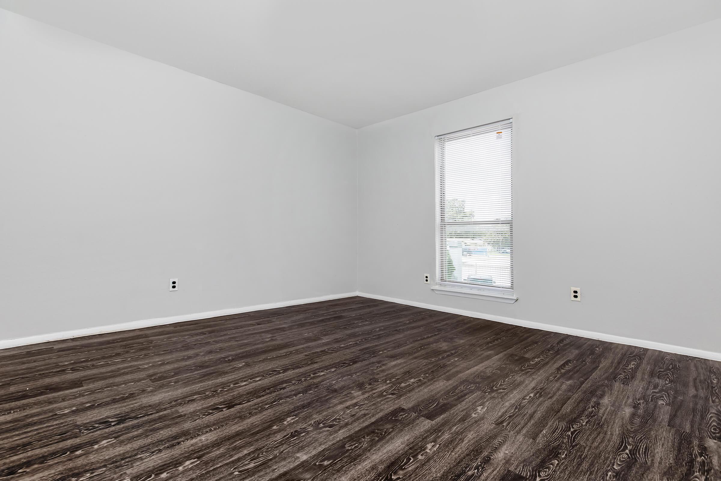 An empty room with gray walls and dark wood-like flooring. The room features a single window with blinds, allowing natural light to enter. There are no furnishings or decorations, giving it a spacious and minimalistic appearance.