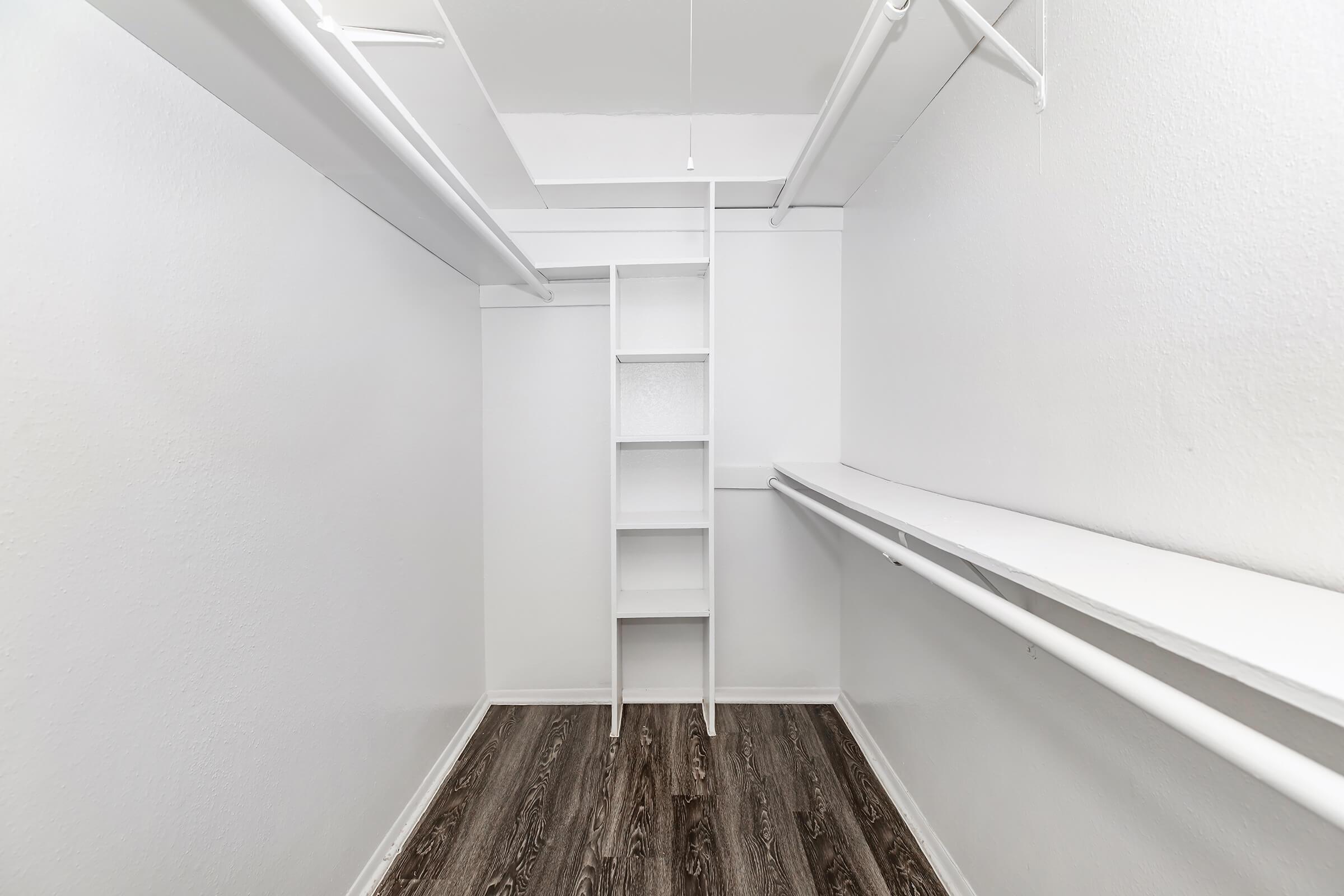 Empty closet featuring white walls, a wooden floor, a built-in shelf unit, and two hanging rods on opposite walls. The minimalistic design creates a spacious feeling, ideal for organizing clothes or storage.