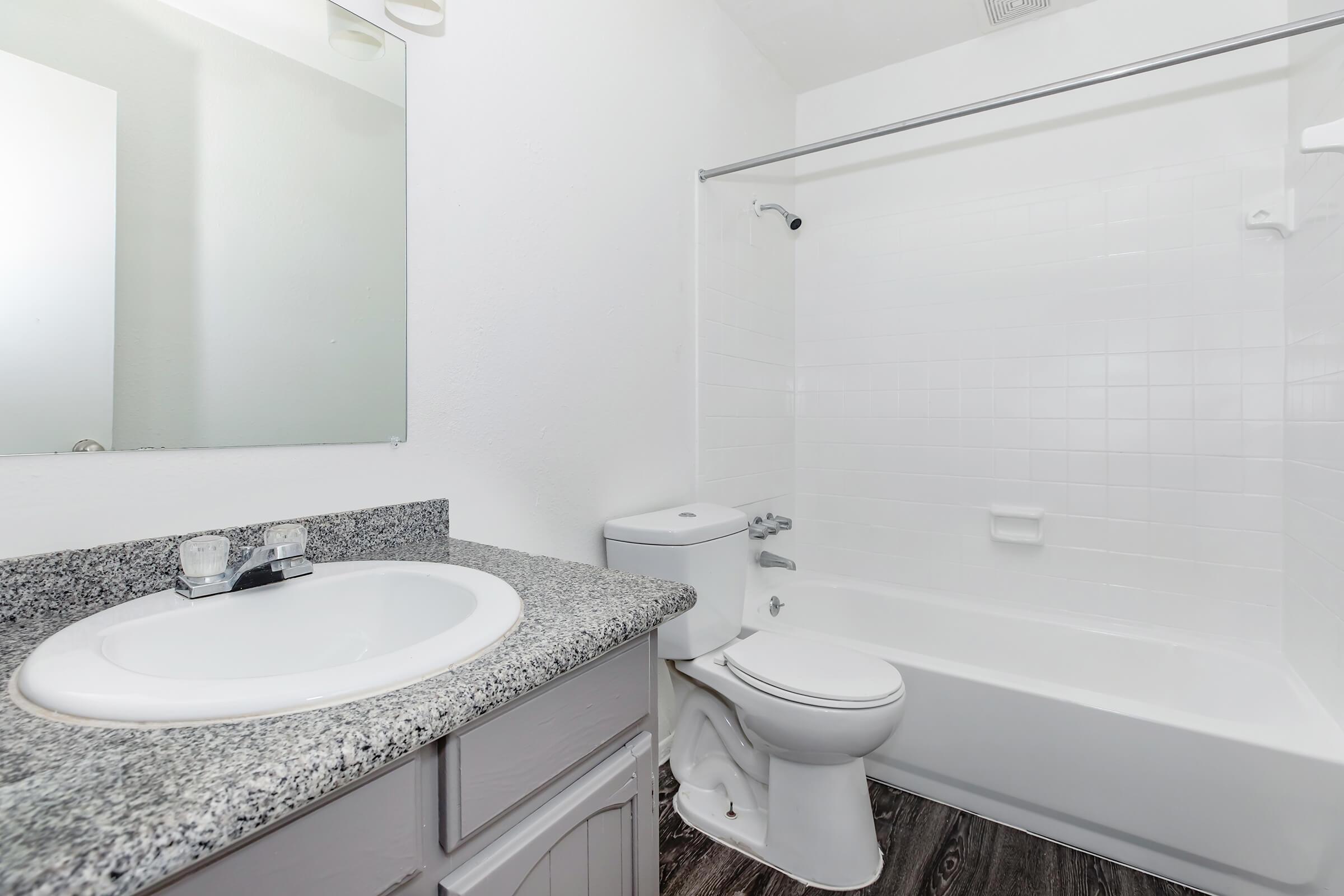 A clean and modern bathroom featuring a white bathtub with a shower curtain, a sink with a granite countertop, a large mirror, and a toilet. The walls are painted white, and the floor has a dark wood-like finish, creating a bright and airy atmosphere.