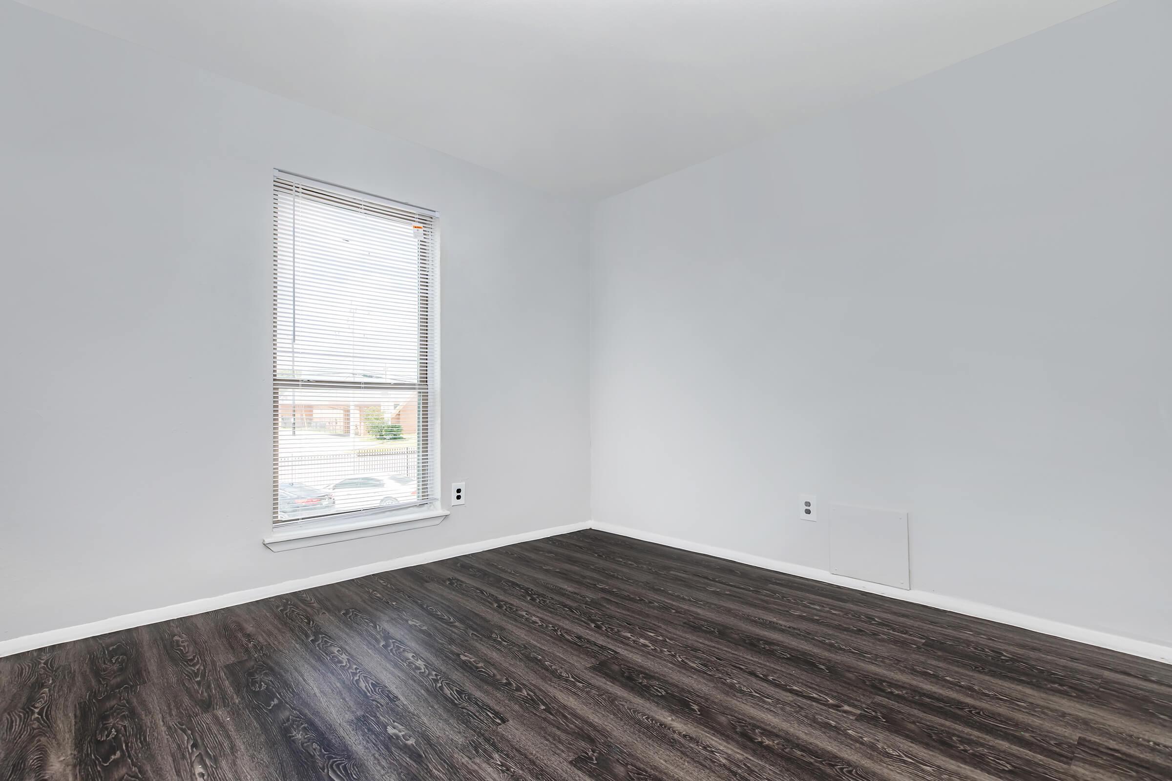 Empty room with light gray walls and dark wood-like laminate flooring. A single window with blinds provides natural light. The room has no furniture, and there is a wall vent near the floor.