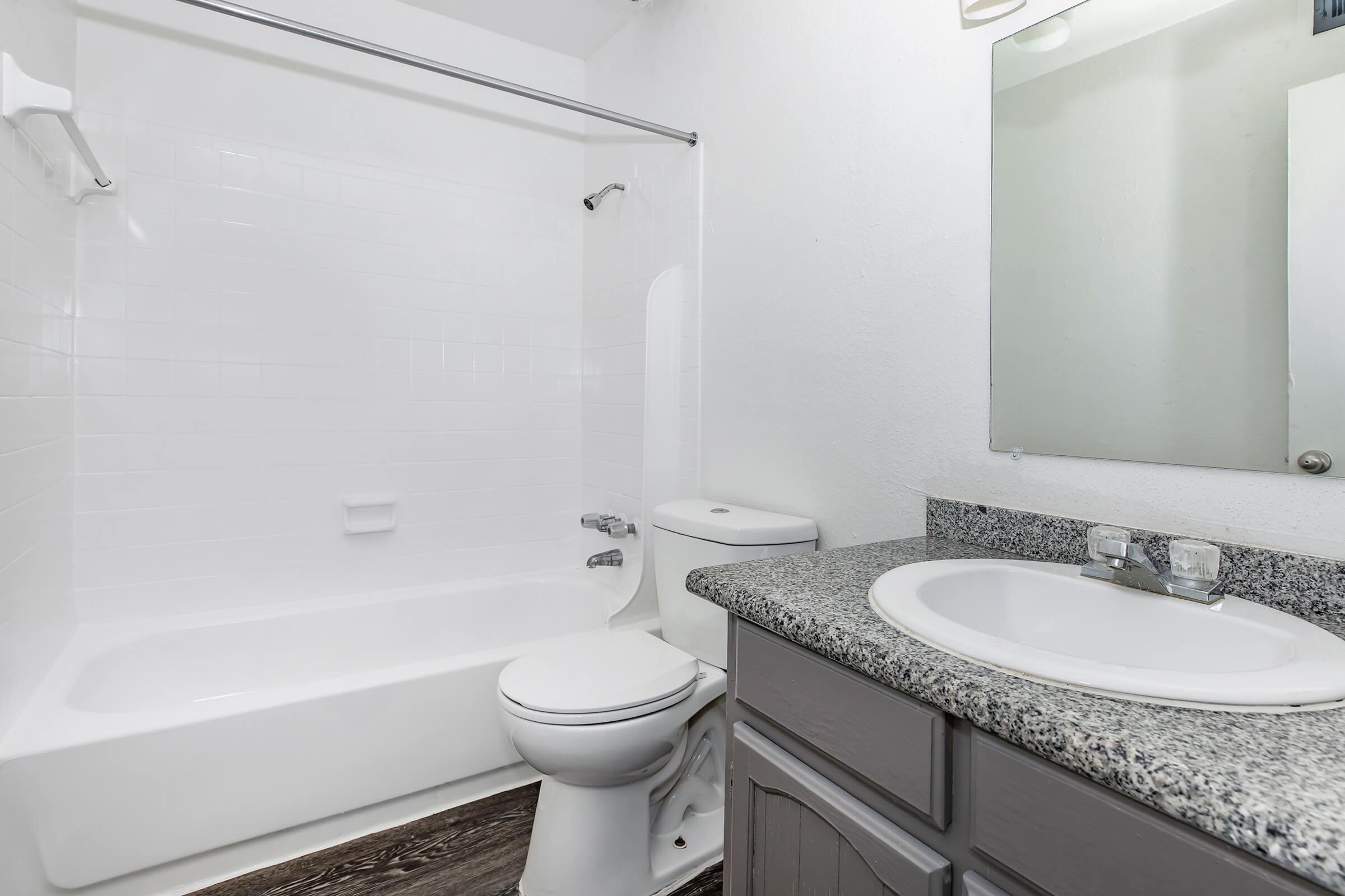 A clean and modern bathroom featuring a white bathtub with a shower, a toilet, and a sink with a granite countertop. The walls are painted white, and there is a large mirror above the sink. The flooring appears to be dark wood.