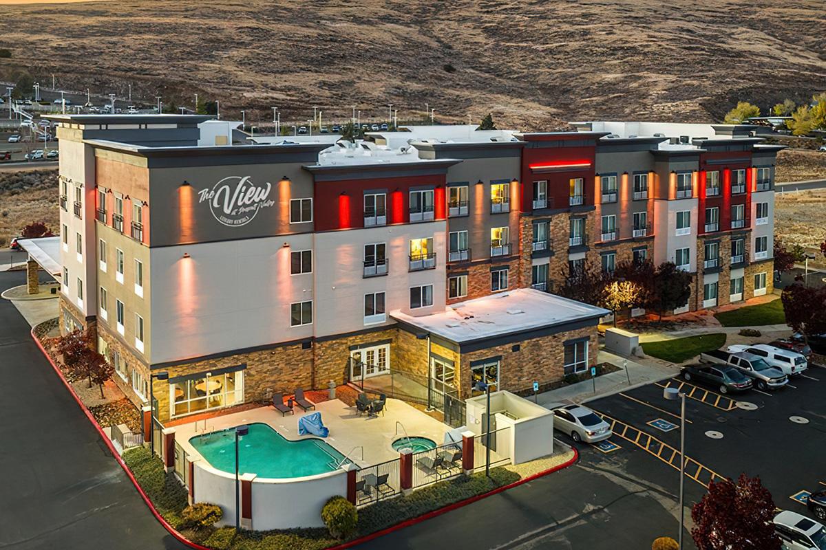 A modern hotel building with a multi-story structure featuring balconies, outdoor lighting, and a swimming pool area. Surrounded by landscaped grounds and parked cars, the setting includes rolling hills in the background and is designed for guests' comfort and relaxation.