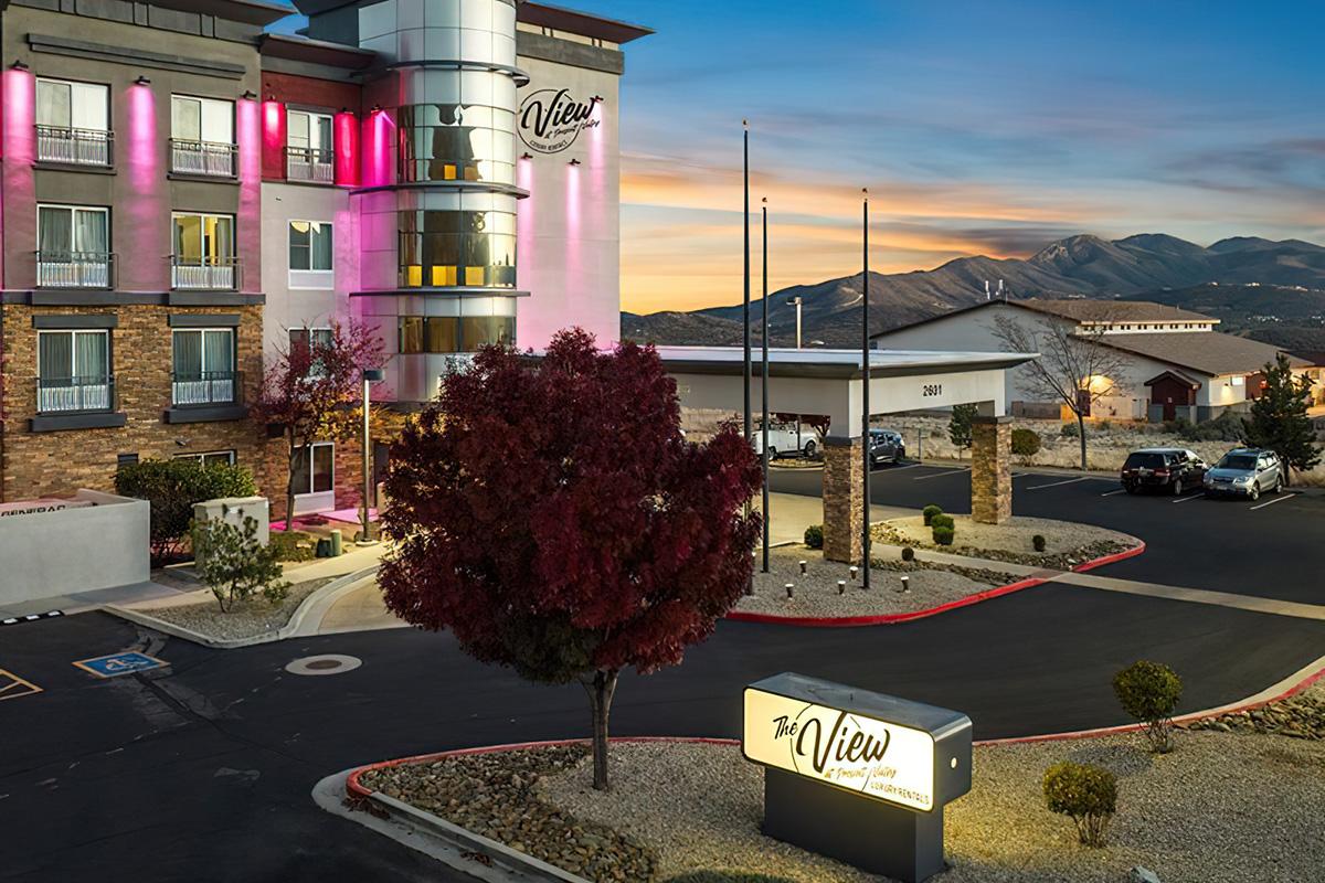 Exterior view of The View hotel at sunset, showcasing a modern building with pink lighting, palm trees, and a welcoming entrance. The surrounding landscape features mountains in the background and well-maintained landscaping in the foreground.