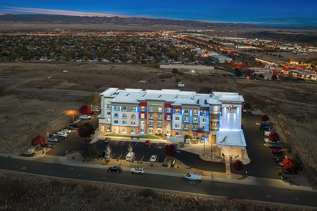 Aerial view of a modern hotel building illuminated at night, surrounded by a parking lot and lined with trees. The hotel features colorful panels and a prominent sign, situated near a town with a sprawling landscape in the background, including roads and buildings.