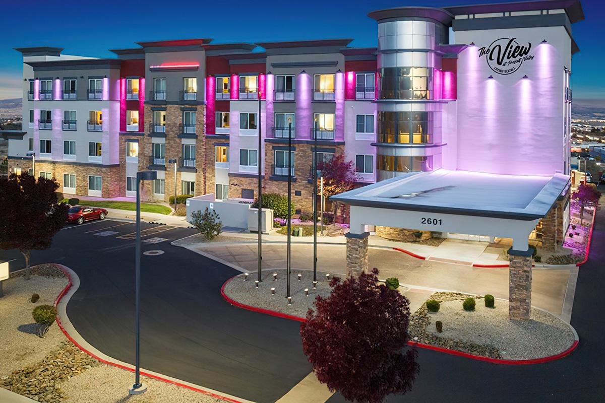 A modern hotel building illuminated with colorful lights at dusk, featuring multiple stories, stone accents, and a covered entrance. The surrounding area includes landscaped plants and a parking lot, with street signage visible.
