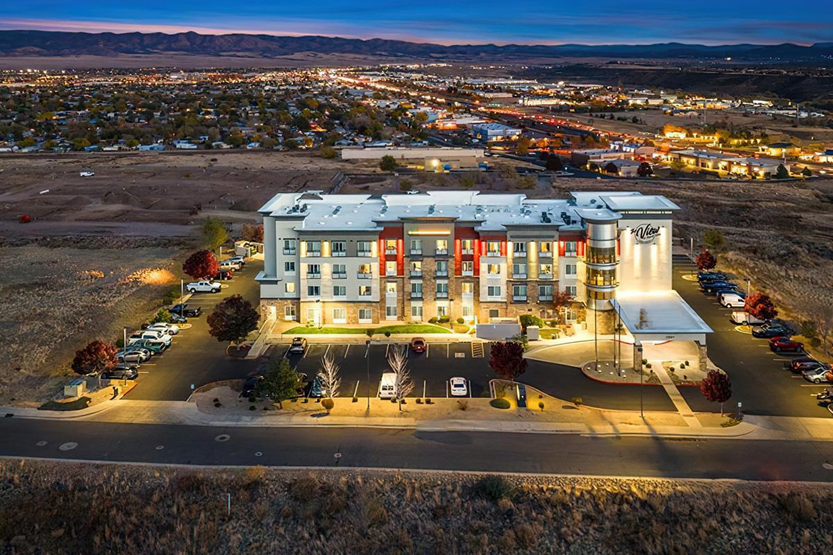 Aerial view of a modern hotel building at dusk, surrounded by parking spaces and illuminated by outdoor lighting. In the background, a sprawling town with roads and hills is visible, creating a scenic urban landscape. The hotel features a distinctive architecture with a cylindrical structure.