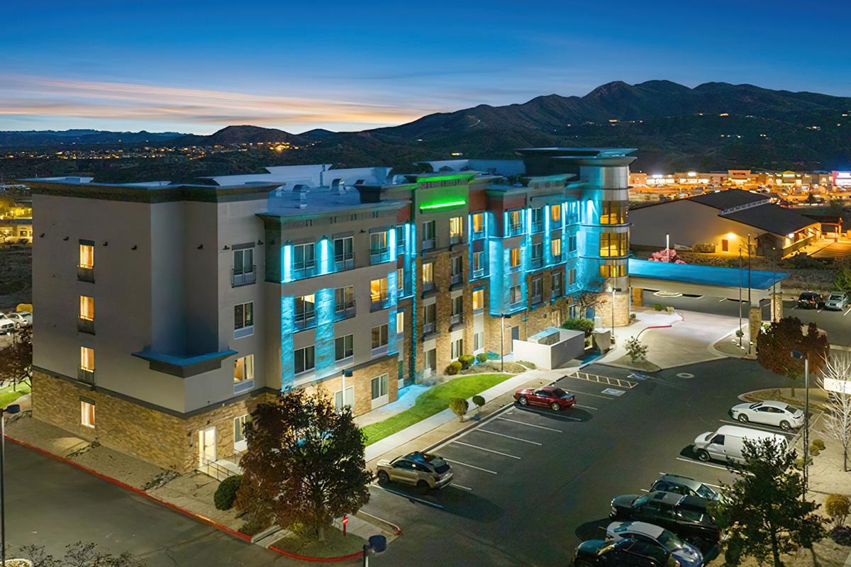 A modern hotel building illuminated with blue lights at dusk, surrounded by a parking lot filled with cars. In the background, rolling hills and city lights are visible, creating a picturesque evening scene.
