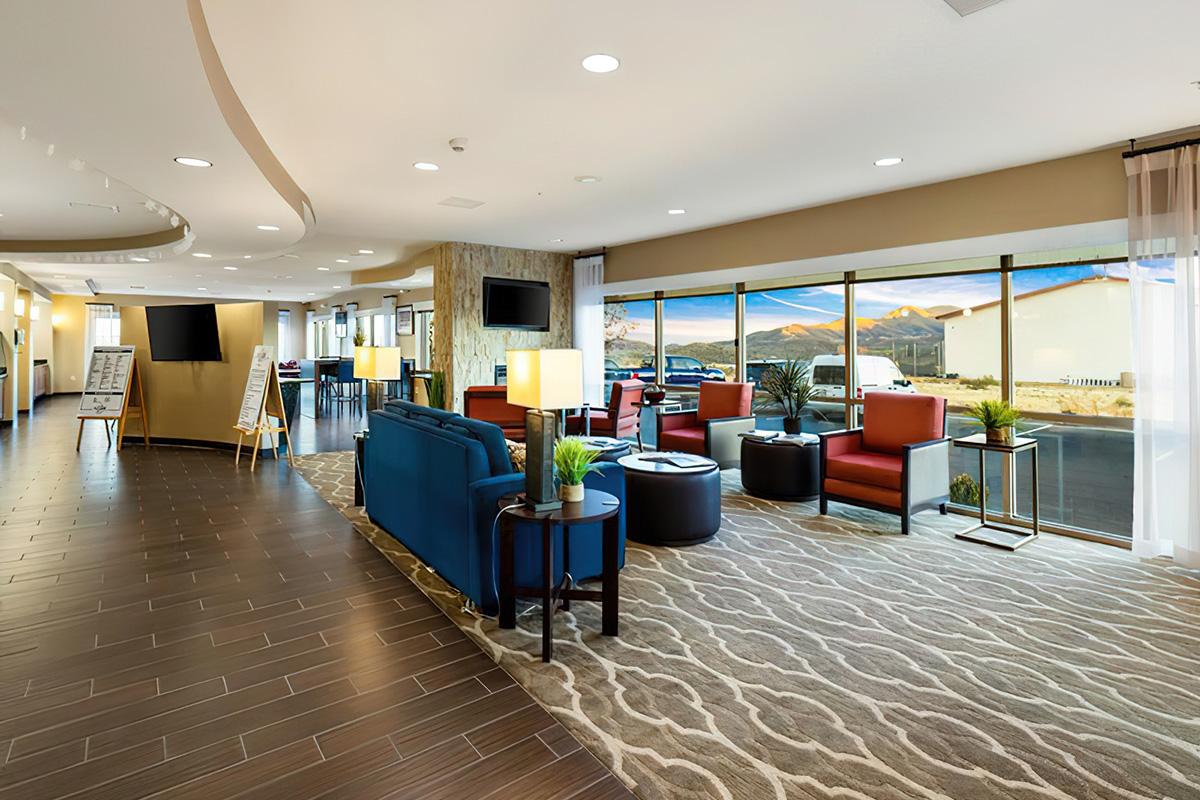 A modern hotel lobby featuring a mix of blue and red seating, stylish tables, and decorative plants. Large windows provide a view of distant mountains, while the floor is adorned with patterned tiles. Informational signs are visible, along with TV screens mounted on the walls.