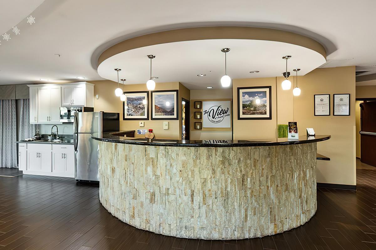 Modern hotel reception area featuring a curved stone-fronted check-in desk, pendant lighting, and framed photographs on the walls. A kitchenette is visible in the background, with white cabinetry, a stainless steel refrigerator, and a welcoming atmosphere. Hardwood floors complete the elegant design.