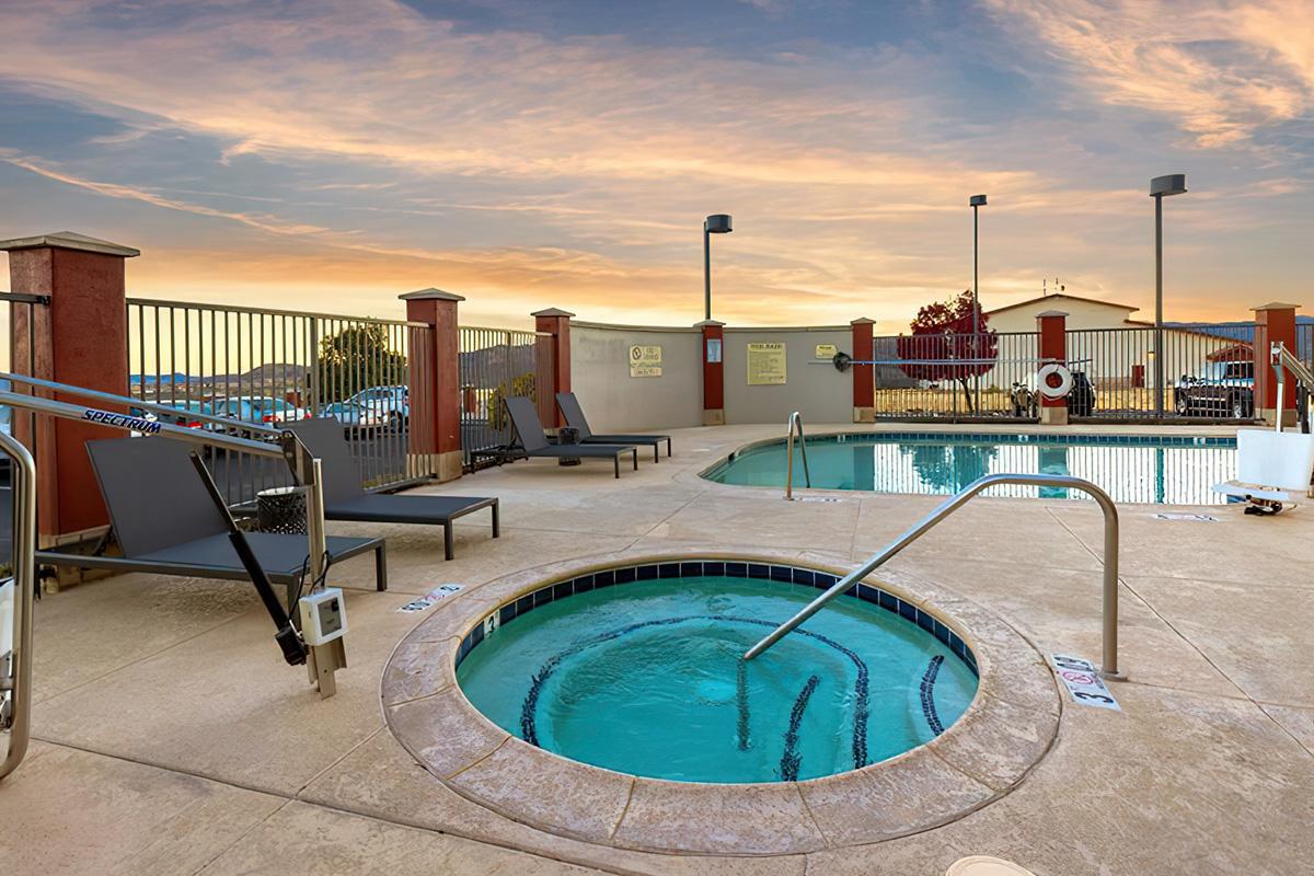 A tranquil outdoor swimming pool area at sunset, featuring a hot tub and lounge chairs. The pool is surrounded by a gated fence and includes signs. Trees and a building are visible in the background, creating a serene atmosphere.