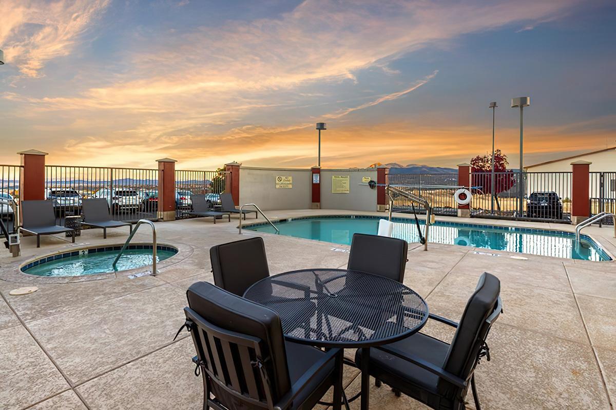 A serene poolside area featuring a swimming pool and a hot tub surrounded by lounge chairs and tables. The scene is enhanced by a beautiful sunset in the background, casting warm colors across the sky. Fences and parked cars are visible in the distance, creating a relaxing atmosphere.