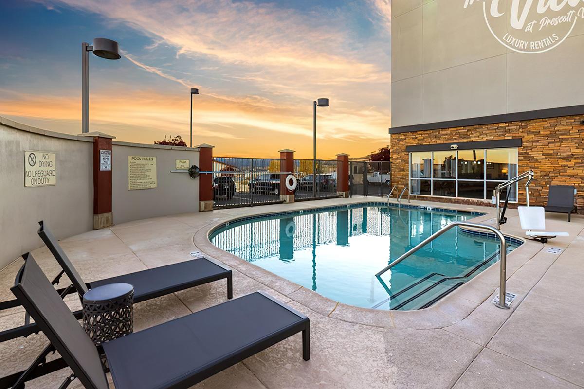 A serene outdoor pool at sunset, featuring two black lounge chairs on the patio. The pool is clear and inviting, surrounded by a fence with a lifebuoy. The building in the background has a modern design, and the sky is painted with warm hues of orange and pink, creating a tranquil atmosphere.