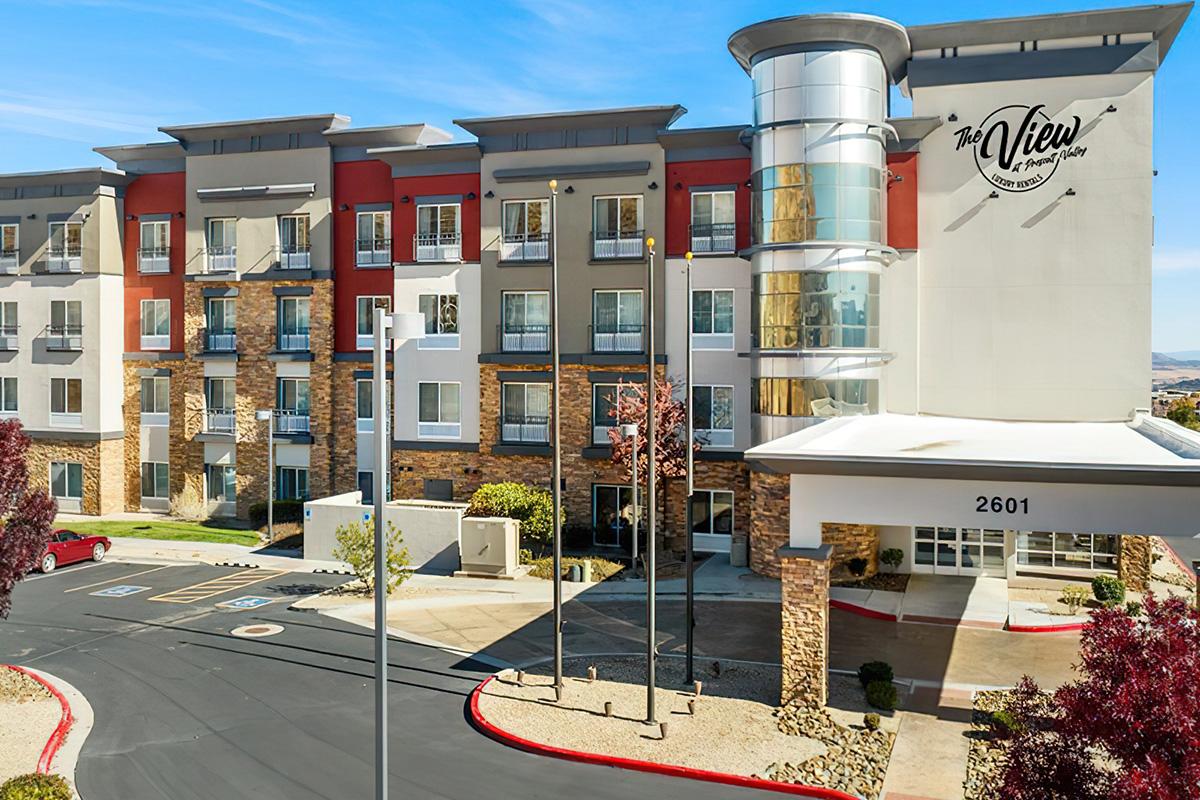 Modern hotel exterior featuring multiple stories with a blend of stone and colorful siding. The entrance is marked by a tall, clear glass structure. Surrounding landscaped areas include trees and parking spaces. Bright, clear sky complements the welcoming atmosphere of the establishment.
