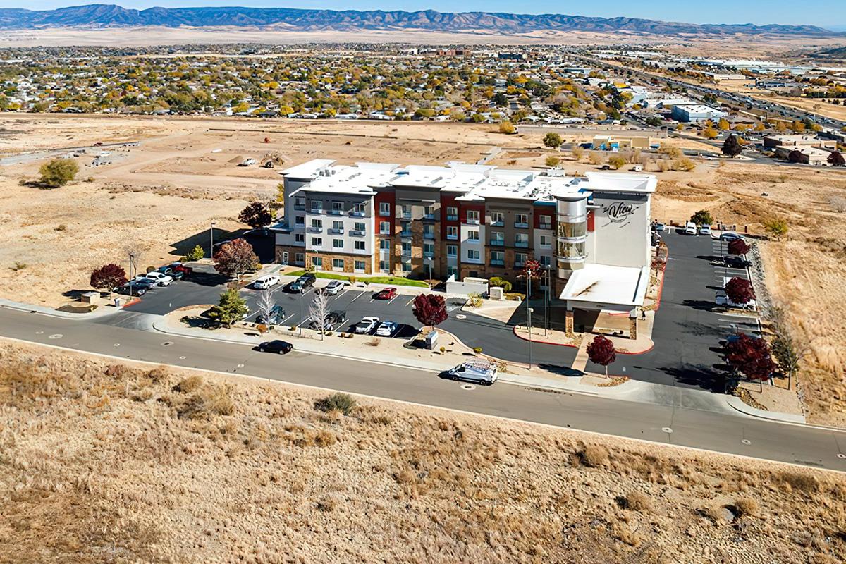 Aerial view of a modern hotel building with multiple stories, surrounded by a parking area. The landscape features arid terrain with sparse vegetation and distant mountains. Nearby, a road runs parallel to the hotel, and other buildings are visible in the background. It's a clear day with a blue sky.
