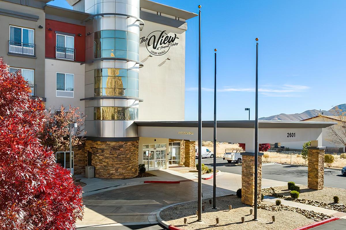 Exterior view of The View hotel featuring a modern entrance with large glass elements and stone accents. Flags are displayed at half-mast in front of the building, surrounded by landscaped areas. Clear blue sky in the background enhances the hotel's vibrant appearance.