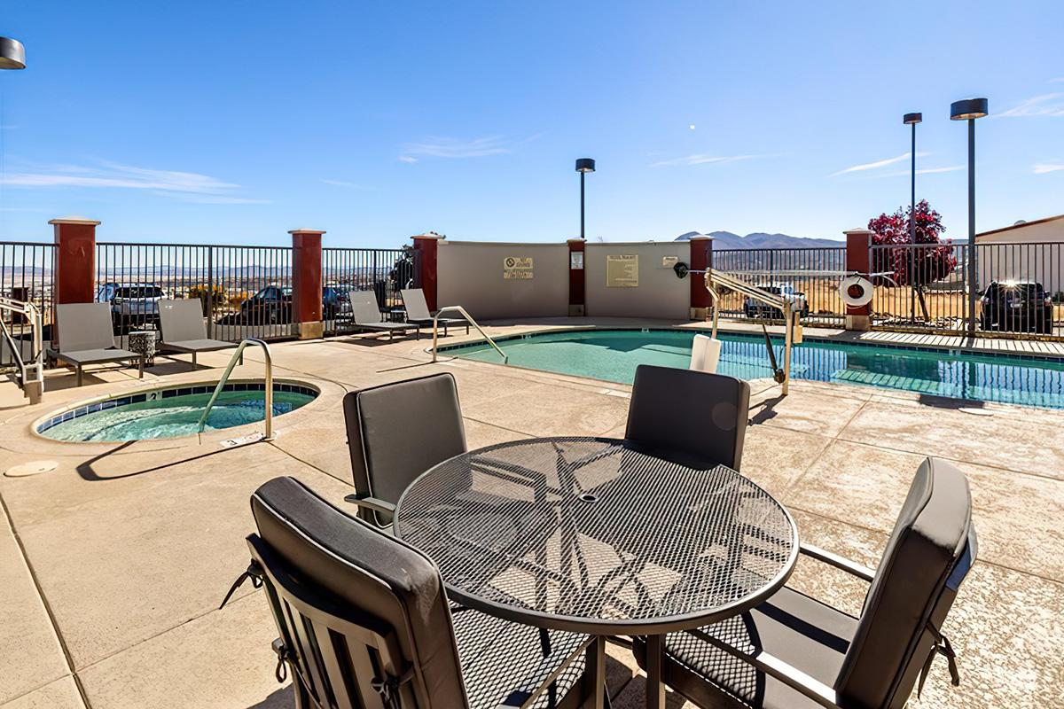 A rooftop swimming pool area featuring a large pool and a hot tub, surrounded by chairs and tables. The scene is sunny, with a clear blue sky and mountains in the background. The pool deck is well-maintained, and there are safety features visible, making it an inviting space for relaxation.