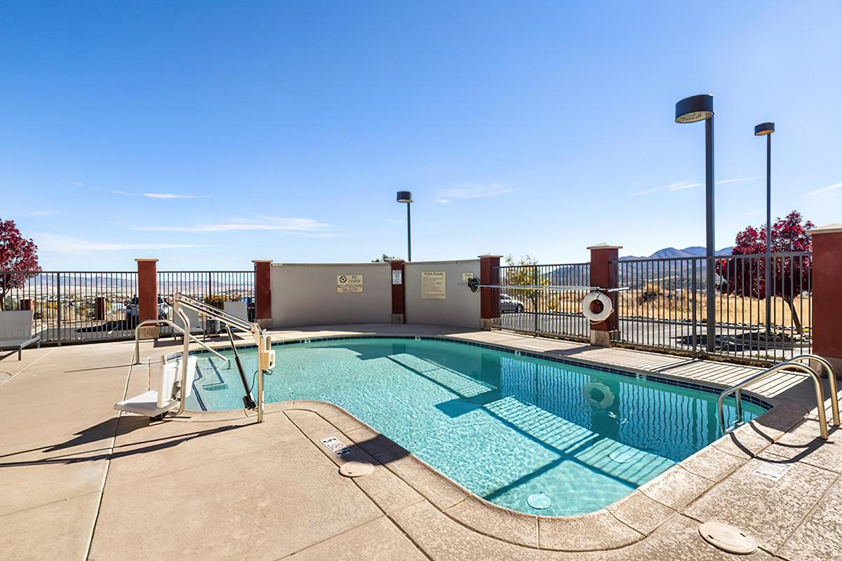 A swimming pool surrounded by a gated patio area, with lounge chairs nearby. The pool features a diving board and is set against a backdrop of clear blue skies and distant mountains. Safety signage is visible, and there are light fixtures overhead.