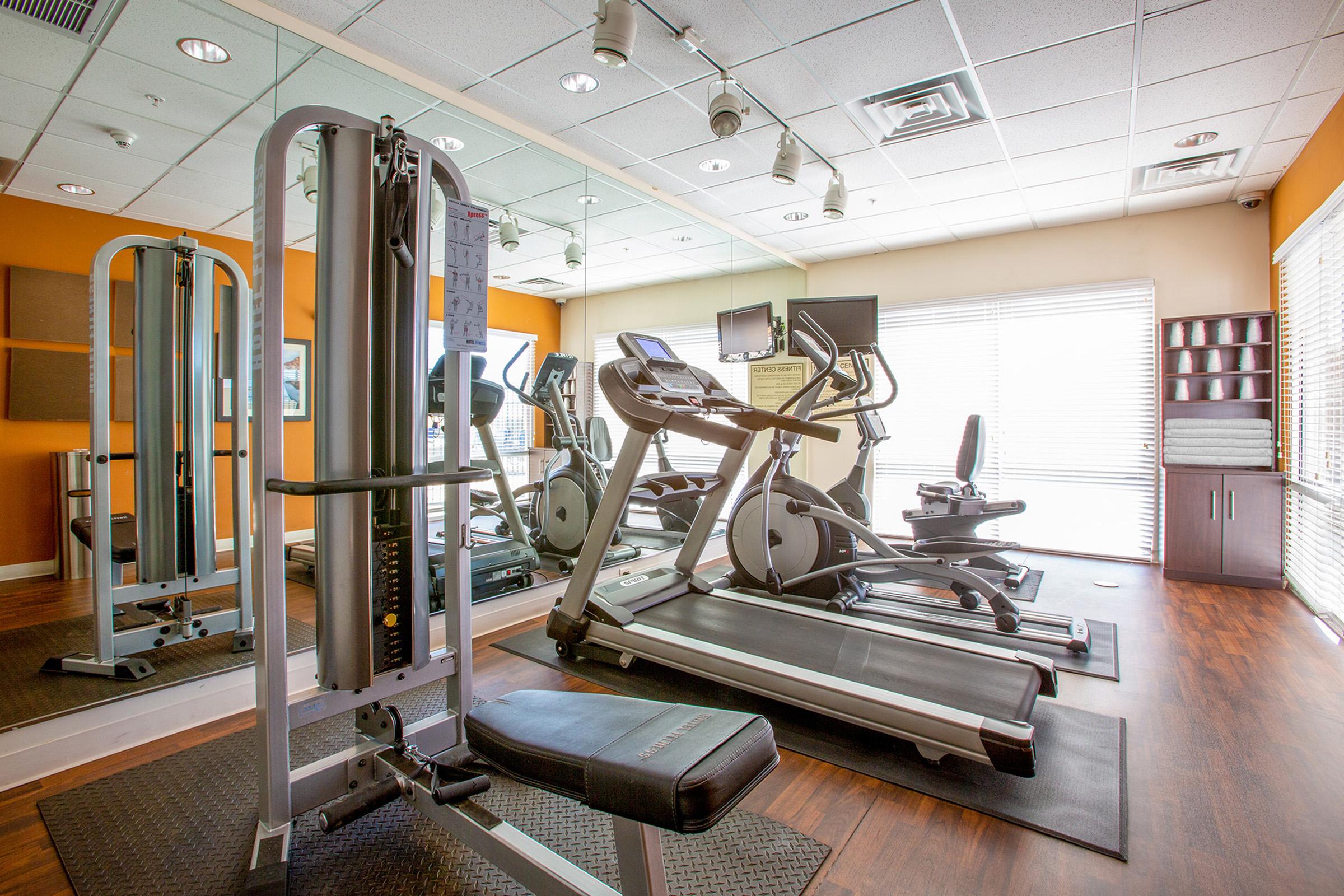 A modern gym interior featuring various exercise equipment, including a weight machine, treadmills, and stationary bicycles. Large windows provide natural light, and mirrors reflect the space, creating an open and inviting atmosphere for workouts.