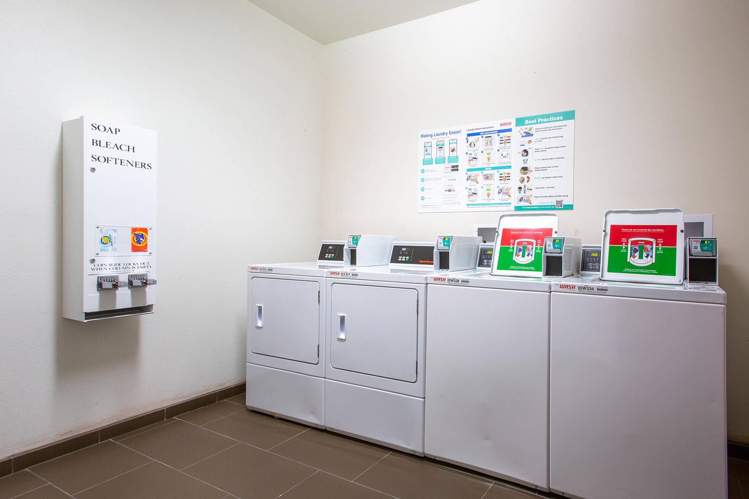 A laundry room featuring three washing machines and a vending machine for soap and bleach. The machines have digital displays, and there is a wall-mounted sign with laundry instructions and best practices. The room has neutral-colored walls and tiled flooring, creating a clean, functional space.