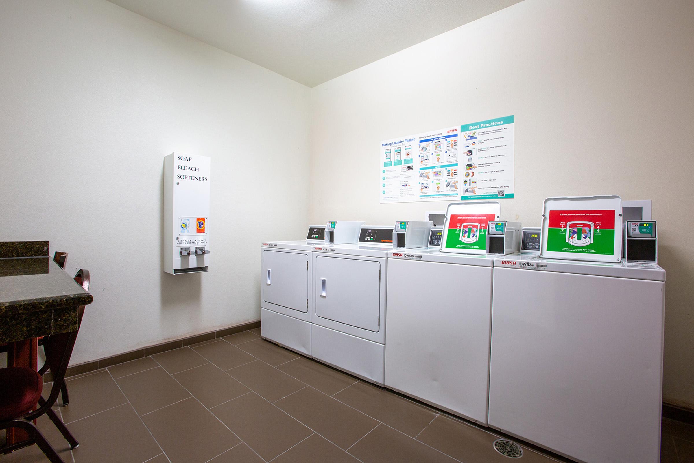 A well-lit laundry room featuring several white washing machines and dryers against a plain wall. To the side, there's a table with chairs and a soap vending machine. A poster above the machines provides laundry instructions and tips. The floor has clean, brown tiles.