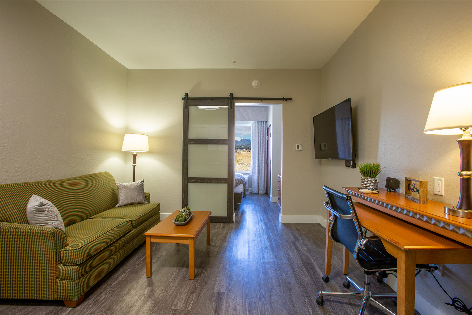 Cozy hotel room featuring a green checkered sofa, a wooden coffee table, a desk with a chair, and a flat-screen TV. The room has neutral walls and hardwood flooring, with a sliding glass door leading to a view of the outdoors. Soft lighting from a lamp creates a warm atmosphere.
