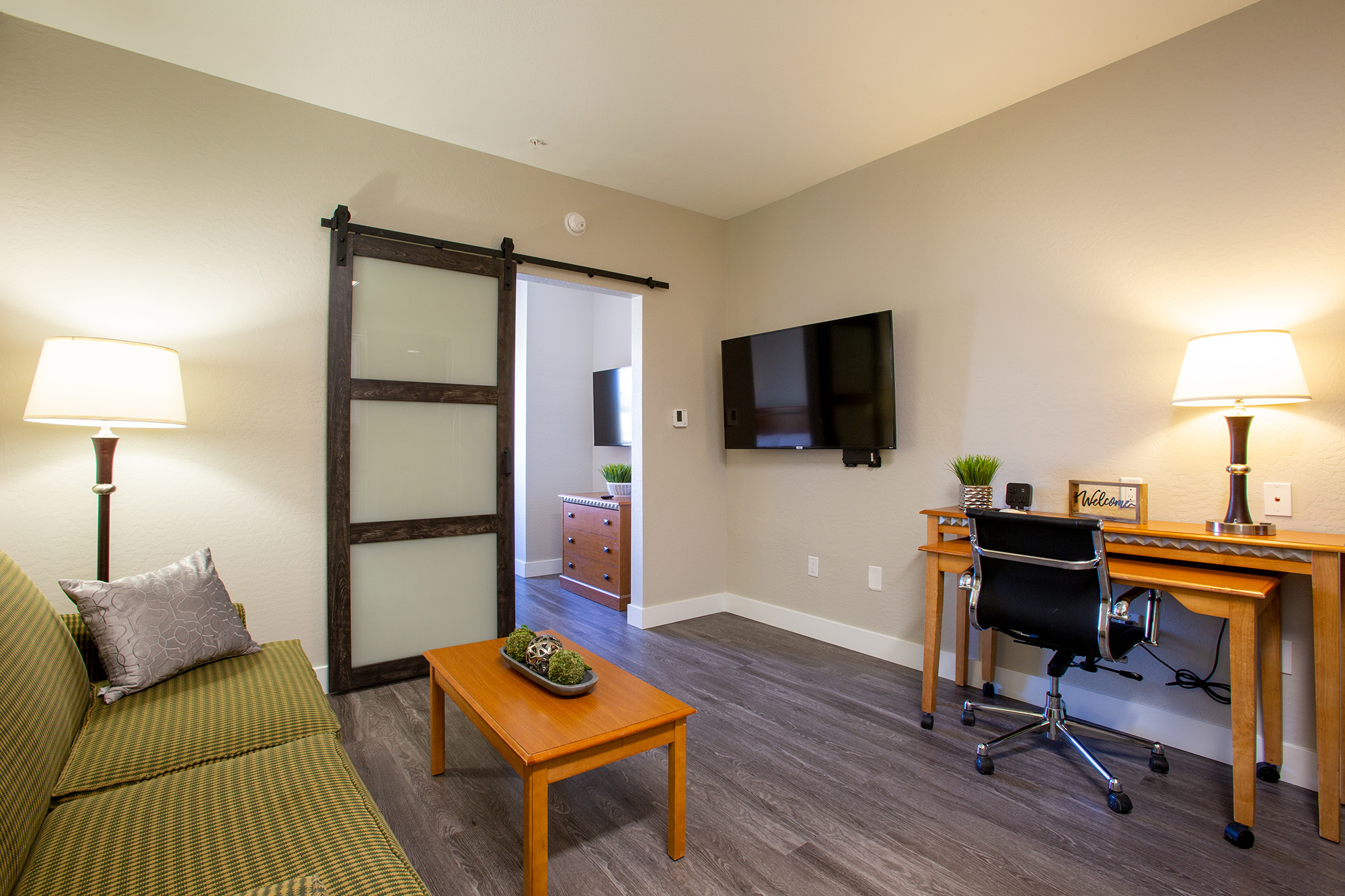 A modern room featuring a sofa with green striped upholstery, a wooden table, and a workspace with a black chair. There are two lamps providing lighting, a television mounted on the wall, and a sliding door leading to another room. The decor is simple and contemporary with potted plants.