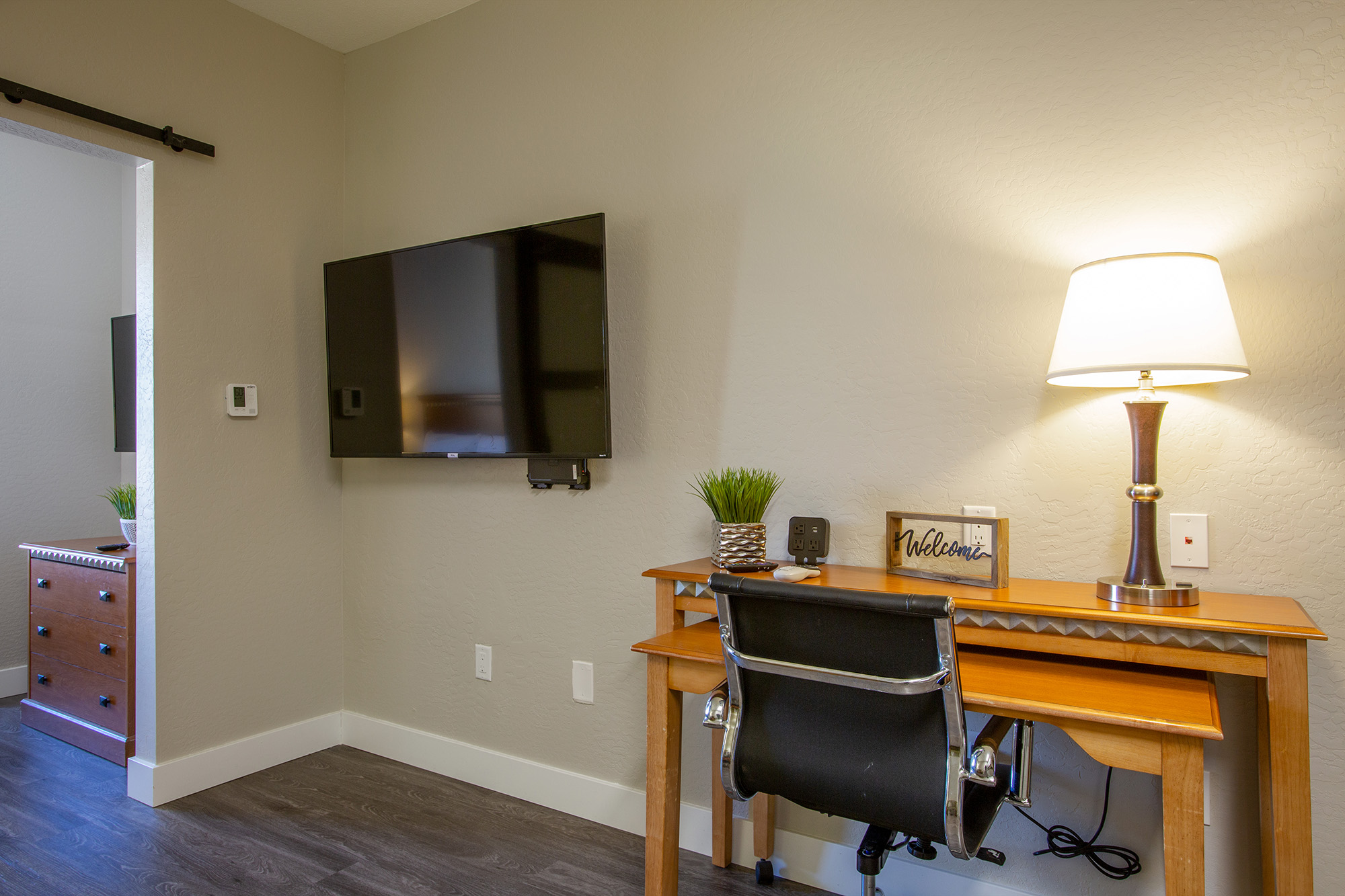 A modern room featuring a wooden desk with a black office chair, positioned near a wall-mounted television. A table lamp sits on the desk, illuminating a decorative sign that reads "Welcome." The walls are painted a light color, and a wooden dresser is visible through an open doorway.