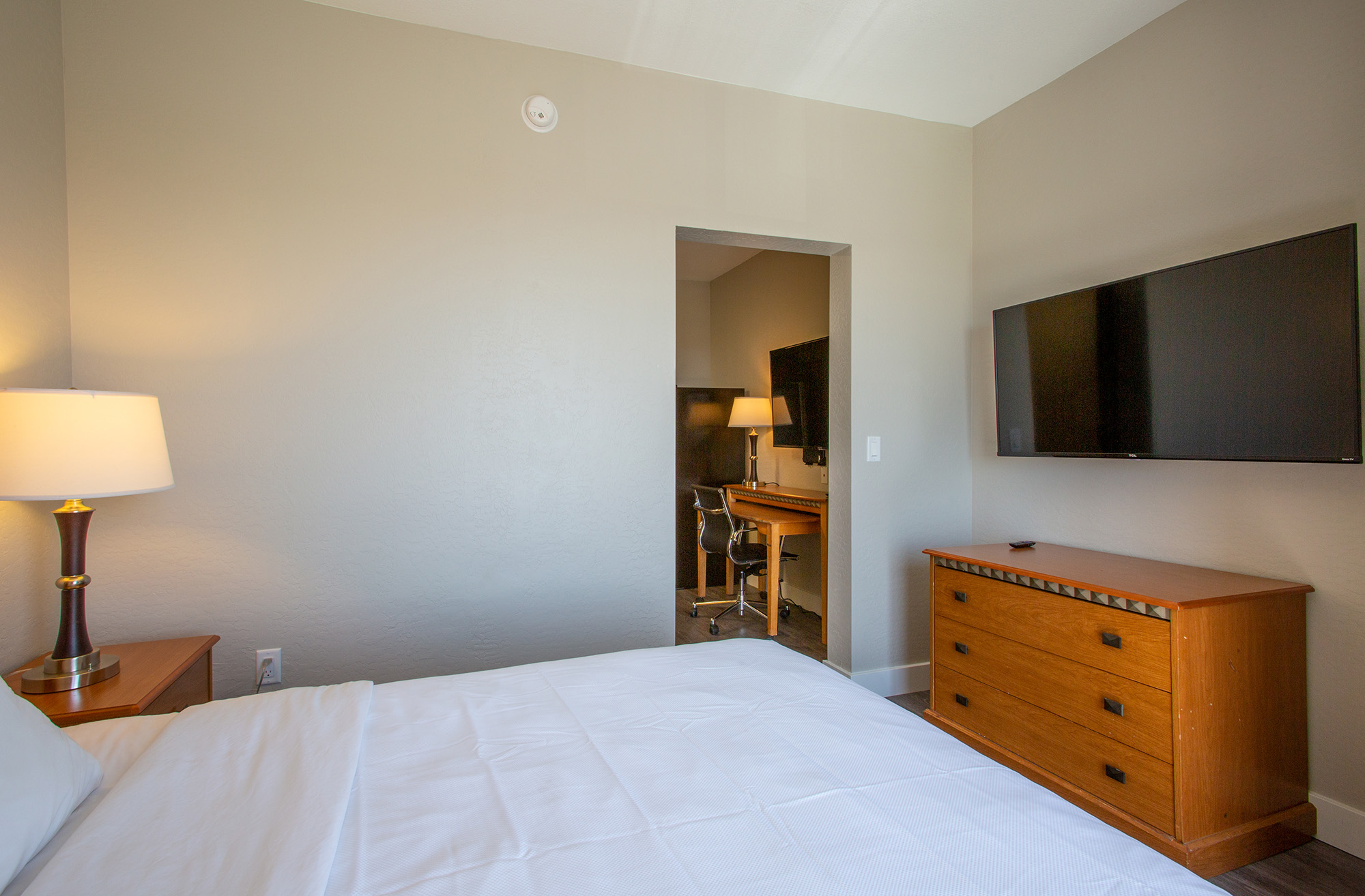 A well-lit hotel room featuring a bed with white bedding, a bedside lamp, a wooden dresser, and a wall-mounted TV. An open doorway leads to a separate workspace with a desk and chair, enhancing the room's functionality and comfort. The walls are painted in a neutral color, creating a calm atmosphere.