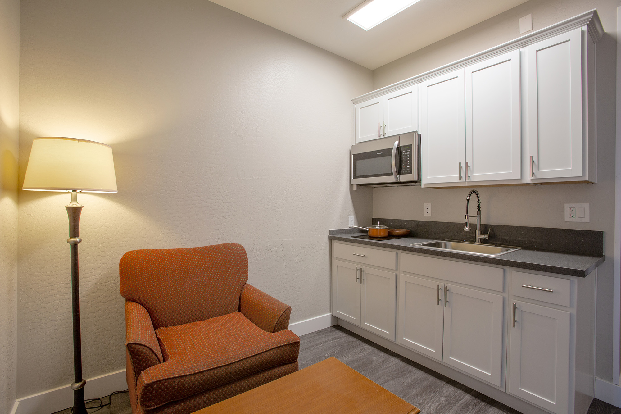 A small kitchenette area featuring a light-colored wall, a sink with a modern faucet, white cabinets, a microwave, and a brown countertop. There's an orange upholstered armchair next to a wooden coffee table and a floor lamp, providing warm lighting. The overall space is tidy and inviting.