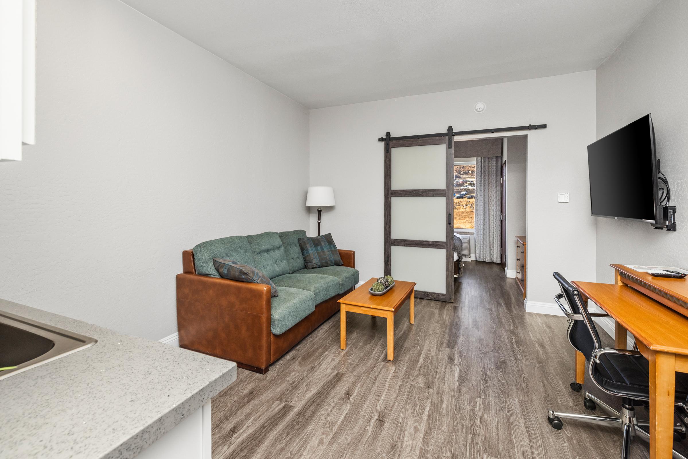 A modern living area featuring a brown couch with decorative pillows, a wooden coffee table, and a flat-screen TV mounted on the wall. A sliding door in the background leads to a bedroom, and there's a desk with a chair nearby. The room has gray walls and wood-like flooring, creating a cozy atmosphere.