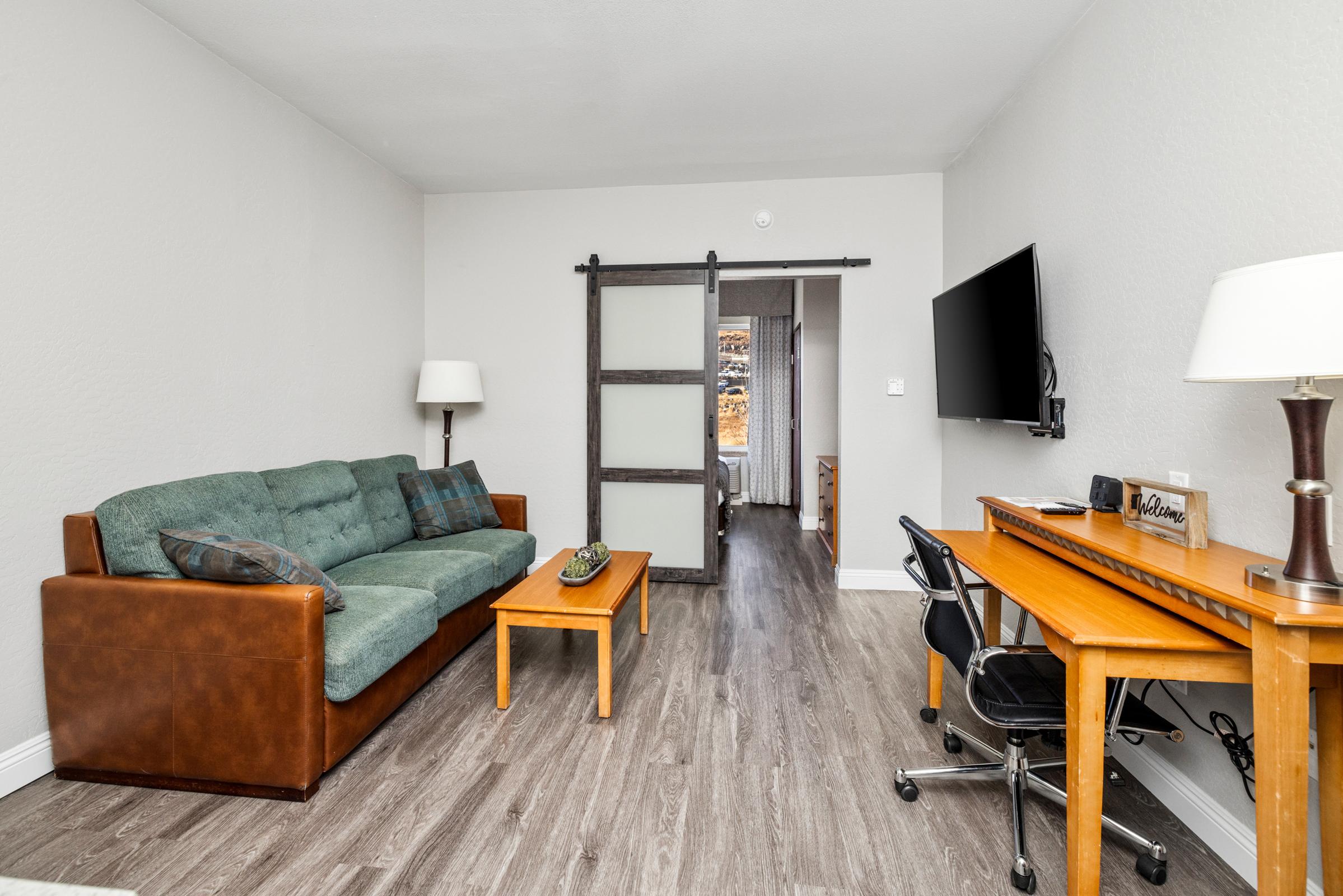 A cozy living room featuring a brown leather sofa with cushions, a wooden coffee table, and a stylish desk with a chair. A flat-screen TV is mounted on the wall, and a sliding door leads to another room. The space has neutral-colored walls and modern flooring, creating a warm and inviting atmosphere.