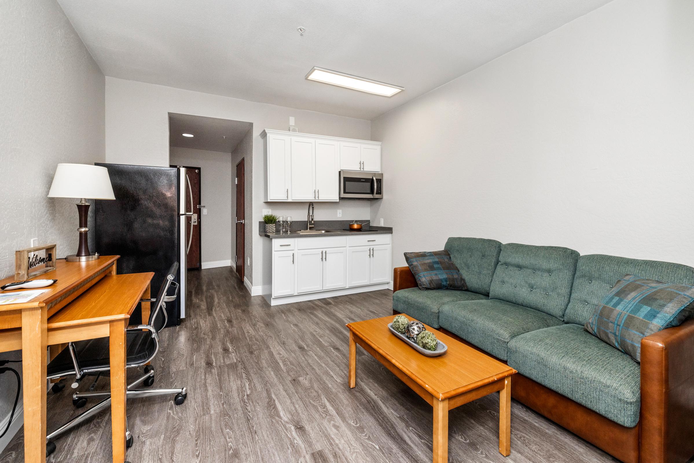 A cozy living space featuring a green sofa, wooden coffee table, and a desk with a chair. The kitchen area includes a refrigerator and microwave, with white cabinetry and a neutral wall color. The floor has a modern wood finish, creating a warm, inviting atmosphere.