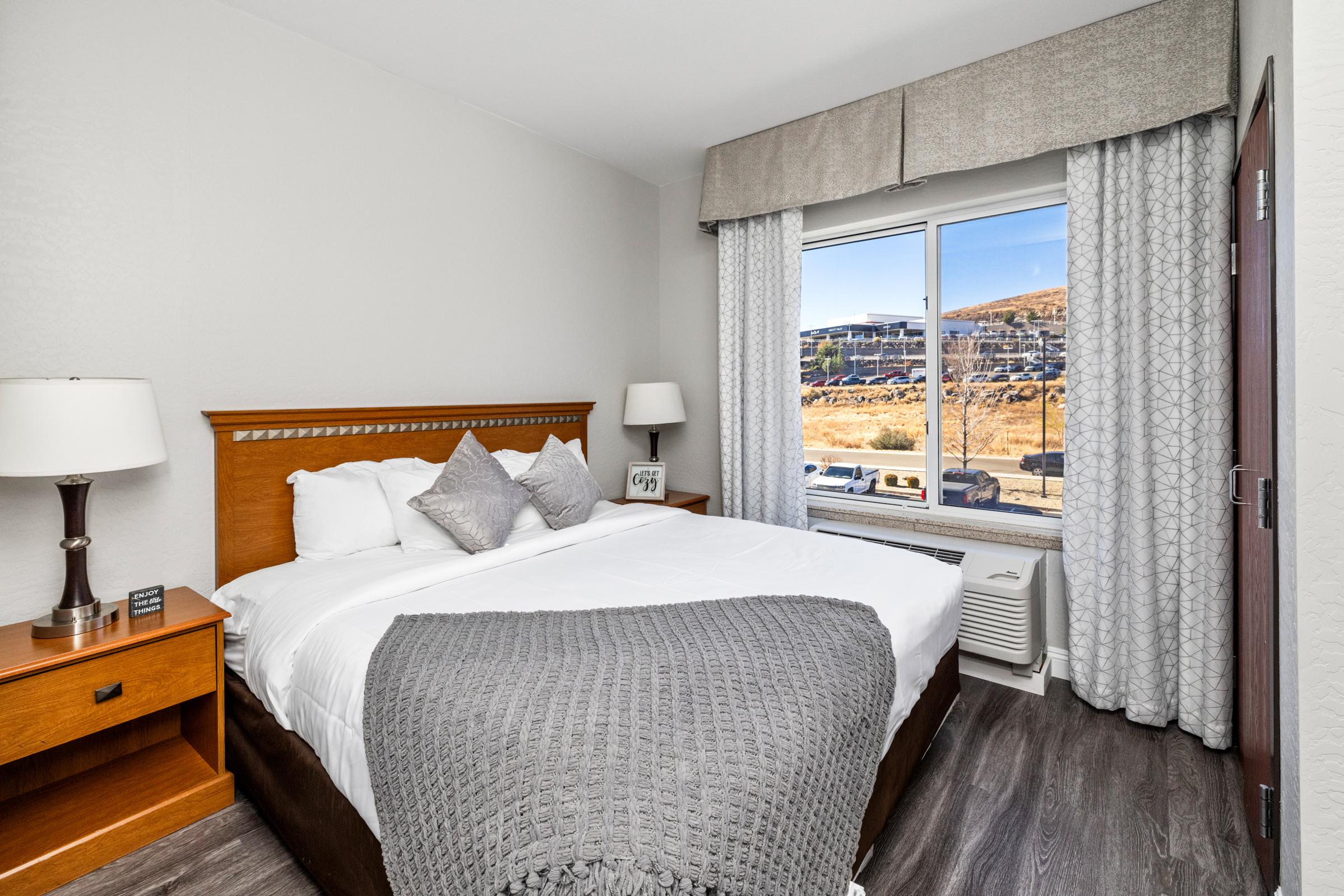 A cozy hotel room featuring a comfortable queen-sized bed with gray linens, two bedside lamps, and a window offering a view of a hillside. The room has modern decor, including a wooden nightstand and an air conditioning unit, creating a welcoming and relaxing atmosphere.