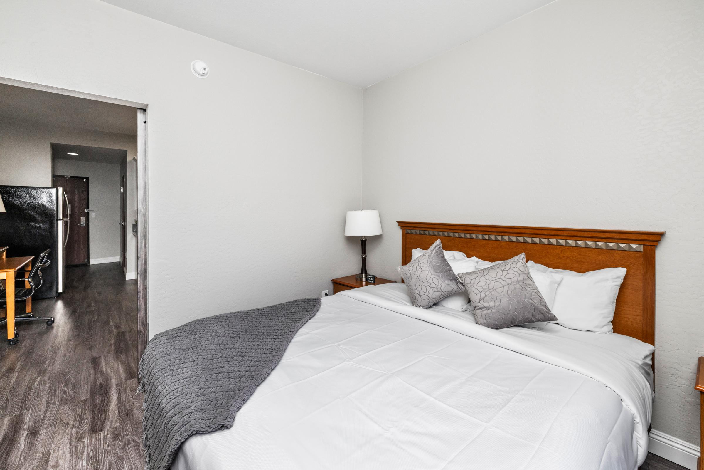 A cozy bedroom featuring a queen-sized bed with white linens and decorative pillows. There's a bedside lamp on a wooden nightstand, and a gray throw blanket drapes across the bed. An open doorway leads to a hallway showing a glimpse of a kitchen area in the background. The walls are painted in a soft gray tone, adding to the inviting atmosphere.