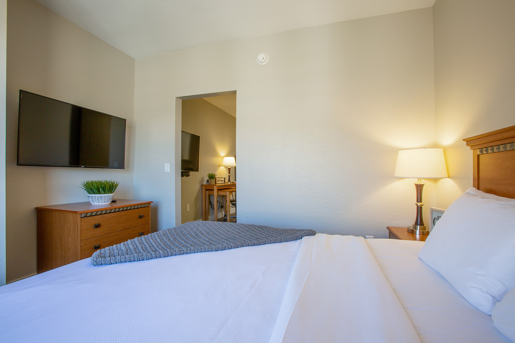 A cozy bedroom featuring a neatly made bed with white linens and a blue throw. There's a wooden nightstand with a lamp and a small plant. A TV mounted on the wall across from the bed, and a doorway leading to another room is visible in the background, illuminating a warm ambiance with soft lighting.