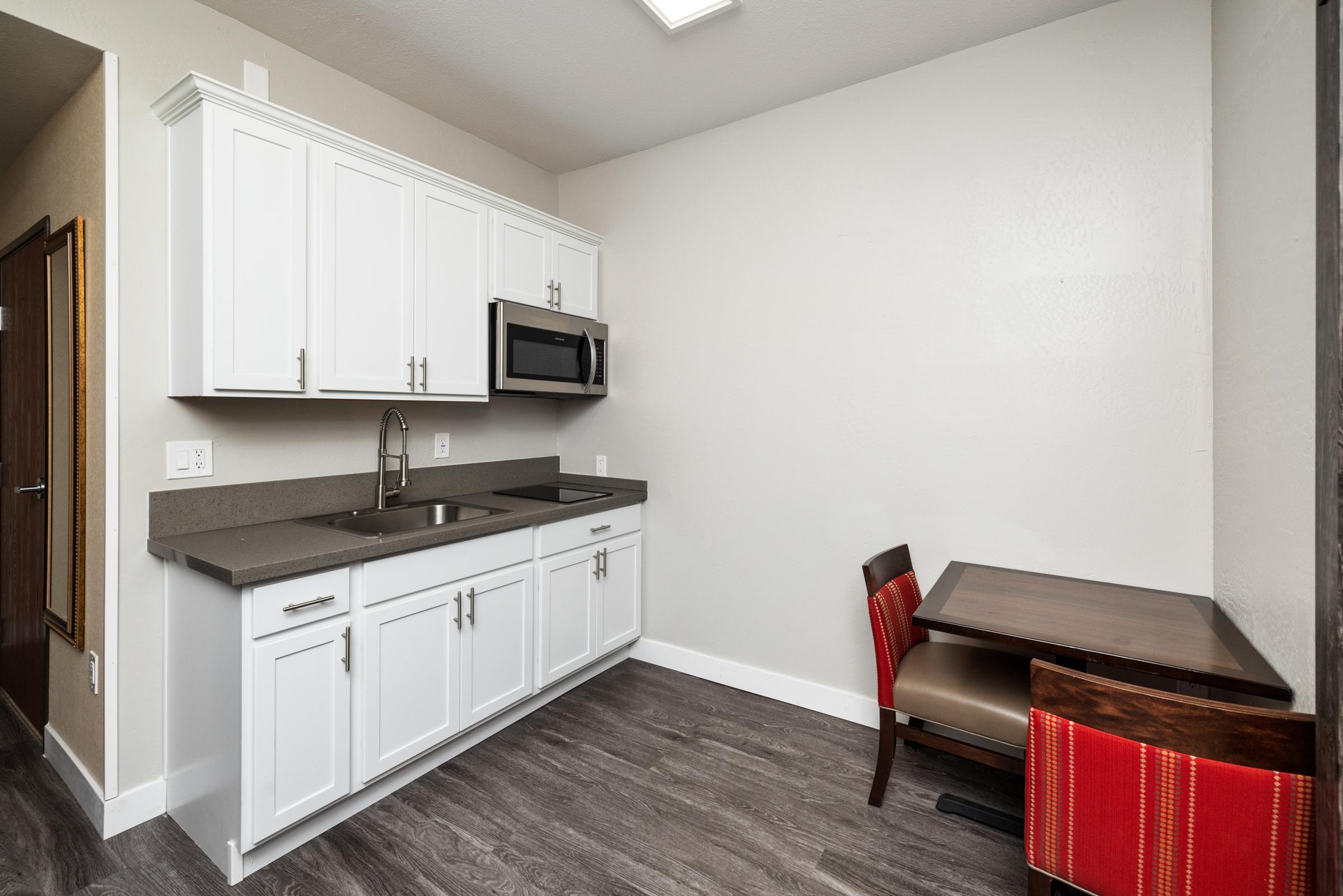 A small kitchen area featuring white cabinetry, a stainless steel microwave, and a sink. There is a compact table with two chairs, one red and one brown, placed against a plain wall. The flooring is a dark wood-like material, contributing to a modern and clean aesthetic.