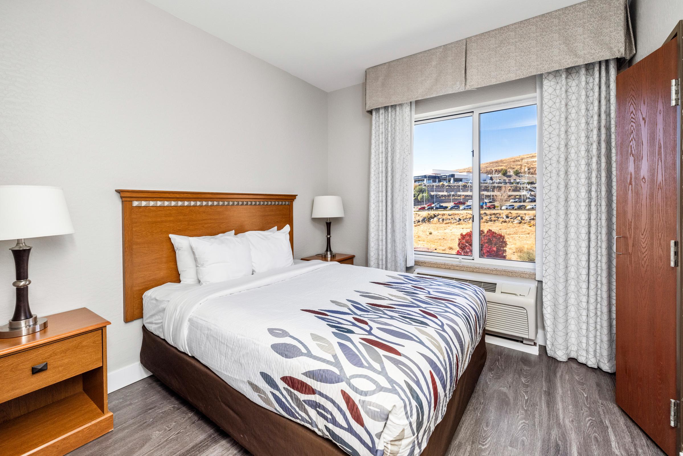 A cozy hotel room featuring a queen bed with white linens, a wooden nightstand with a lamp, and a window showcasing an outdoor view. The walls are painted in neutral tones, and there is a dresser against one wall. Natural light enters the room, creating a welcoming atmosphere.