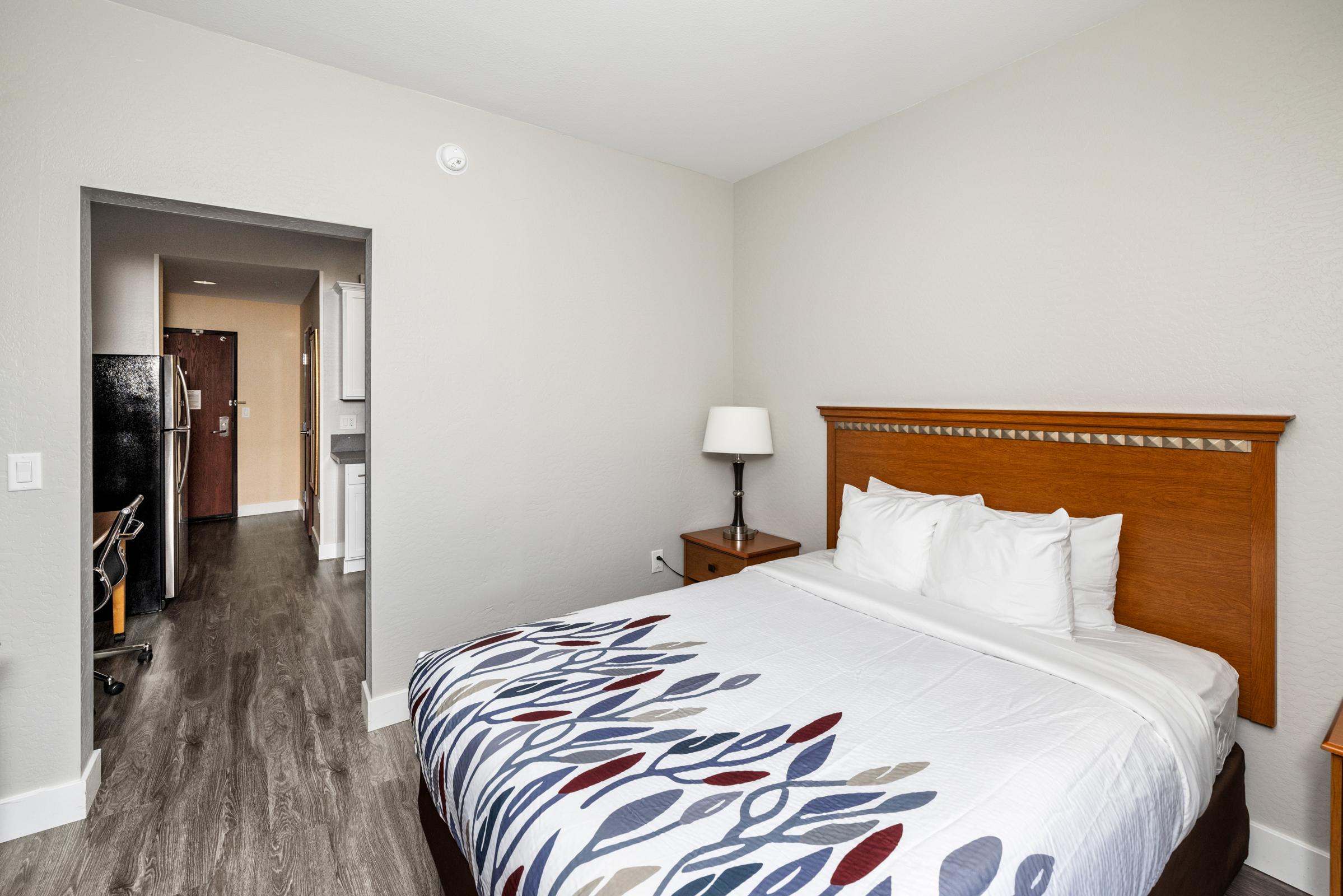 A cozy hotel room featuring a queen-size bed with a patterned comforter, a bedside table with a lamp, and a spacious layout. The room has light-colored walls and wooden flooring. A doorway leads to a hallway with another room visible in the background.