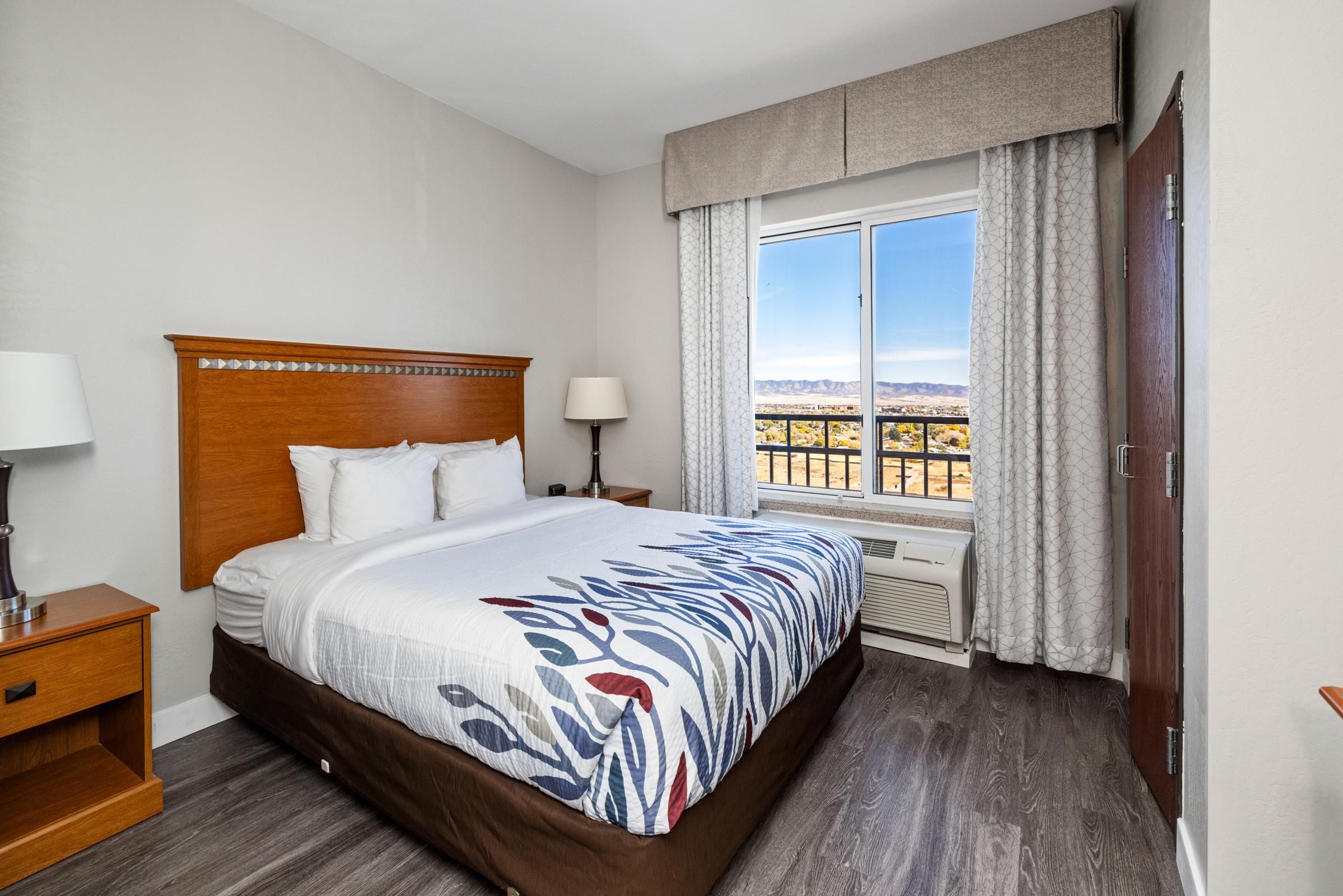 A cozy hotel room featuring a double bed with a decorative headboard, a nightstand with a lamp, and a large window showcasing a scenic view. The room has light-colored walls, wooden flooring, and beige curtains, creating a warm and inviting atmosphere.