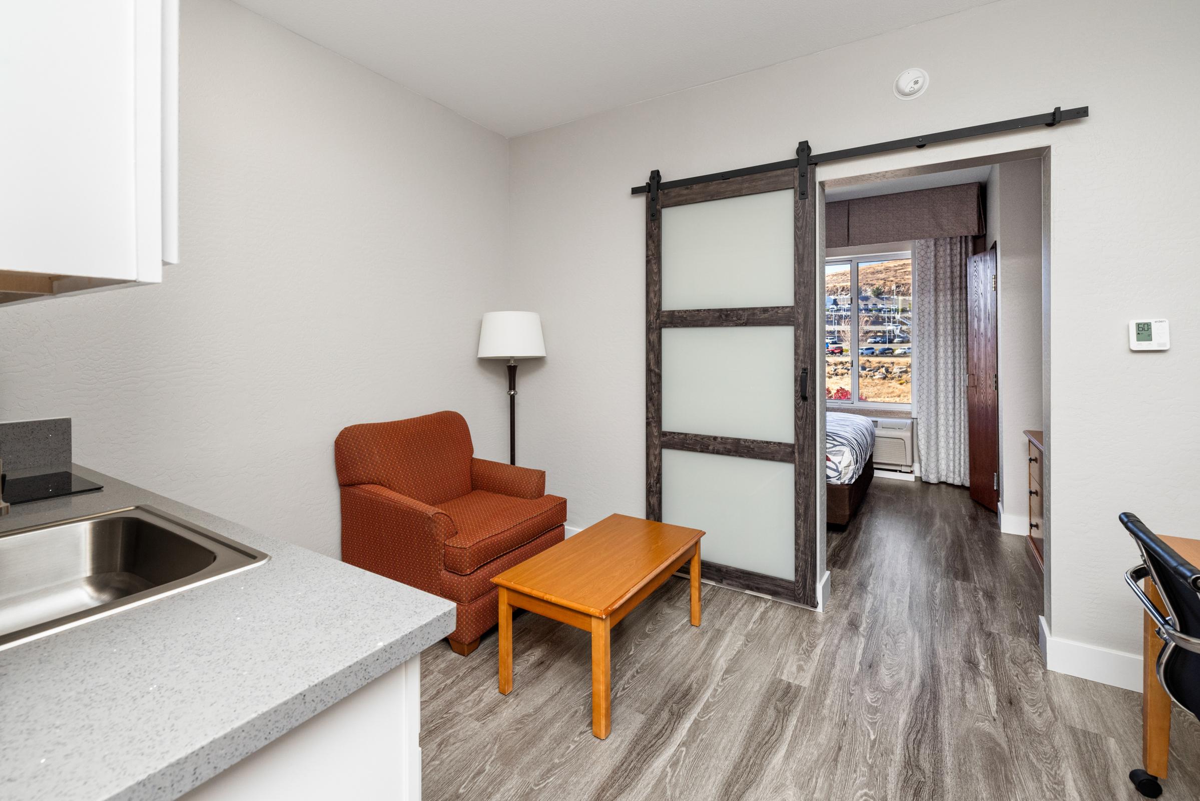 Interior view of a small hotel room featuring a kitchenette with a sink, a cozy orange armchair, and a wooden coffee table. A sliding door leads to a bedroom area visible in the background, with natural light coming through the window.
