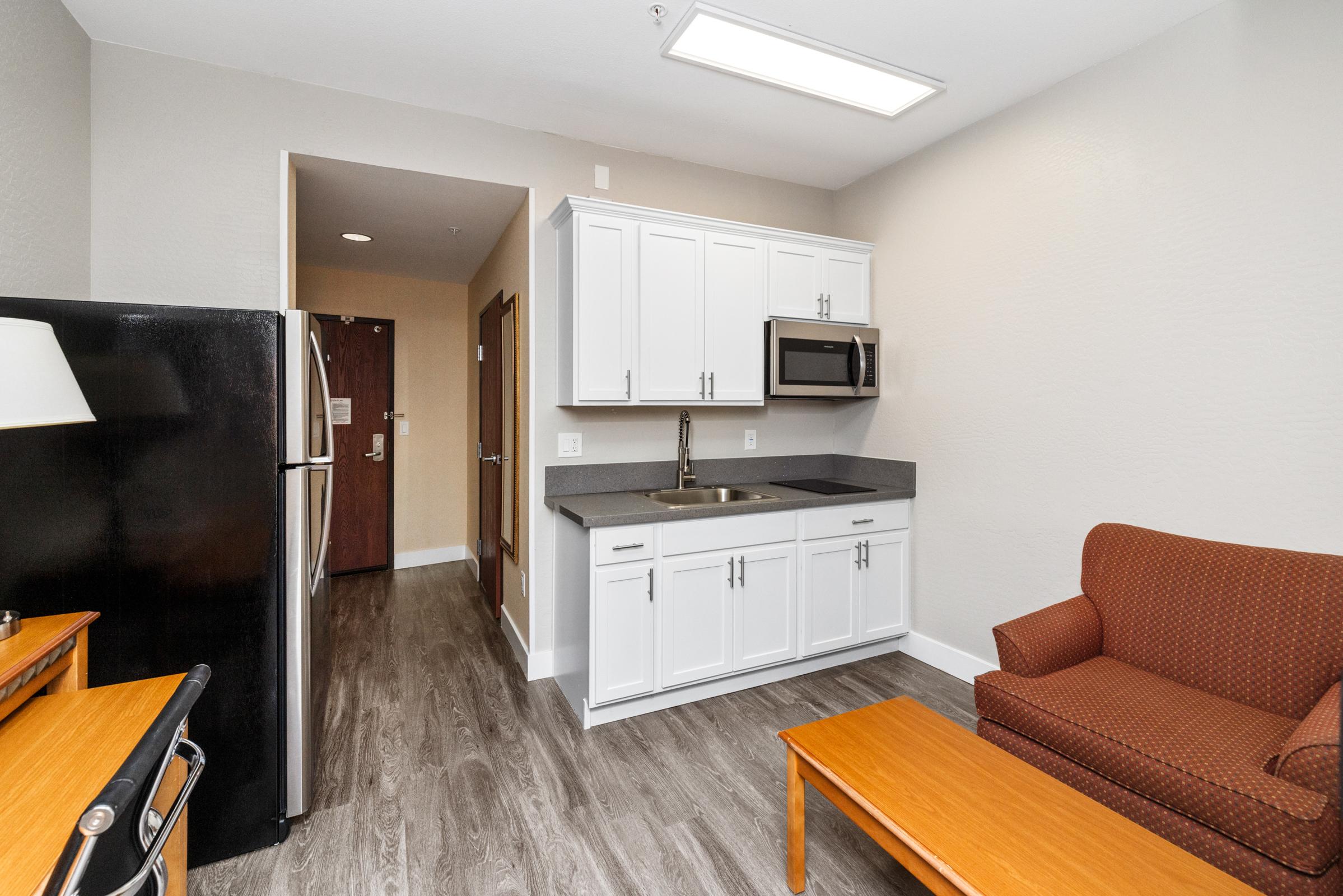 A furnished kitchenette area featuring white cabinets, a sink, and a microwave. There is a black refrigerator, a wooden coffee table, and a brown upholstered sofa. The room has light-colored walls and laminate flooring, with a door leading to another area in the background.